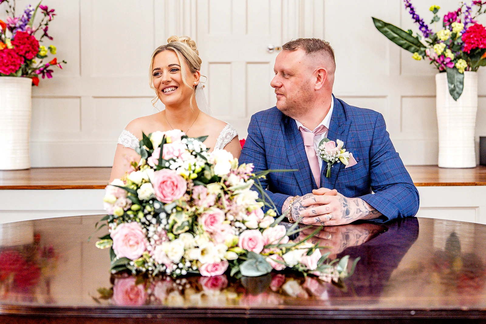 A smiling bride and groom sitting together at a polished table during their wedding ceremony in a light panelled room, with a large floral arrangement in the foreground.