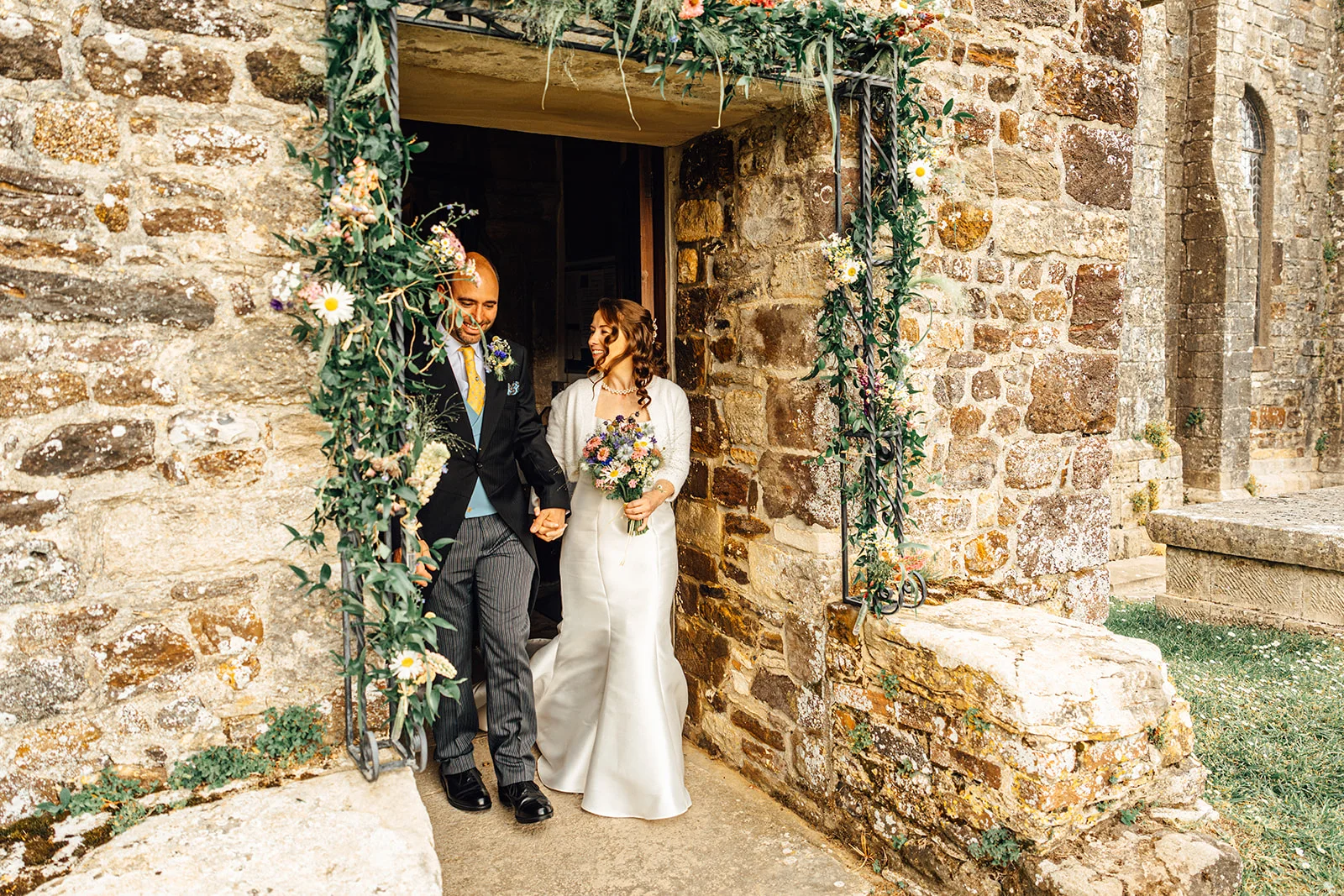 A newlywed couple happily walk hand in hand out of a stone church doorway adorned with a lush floral arch.