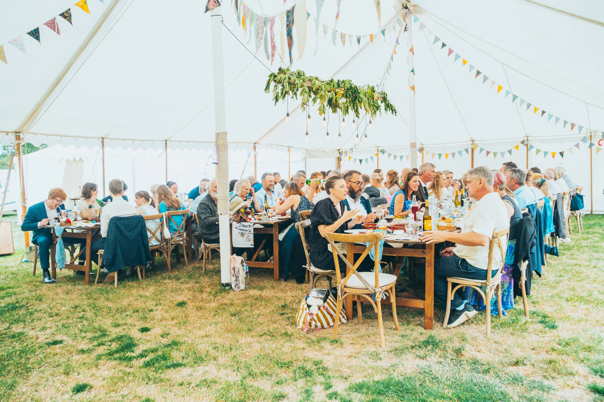 Wedding guests enjoy a relaxed wedding breakfast seated at long wooden banquet tables inside a bright marquee decorated with colourful bunting.