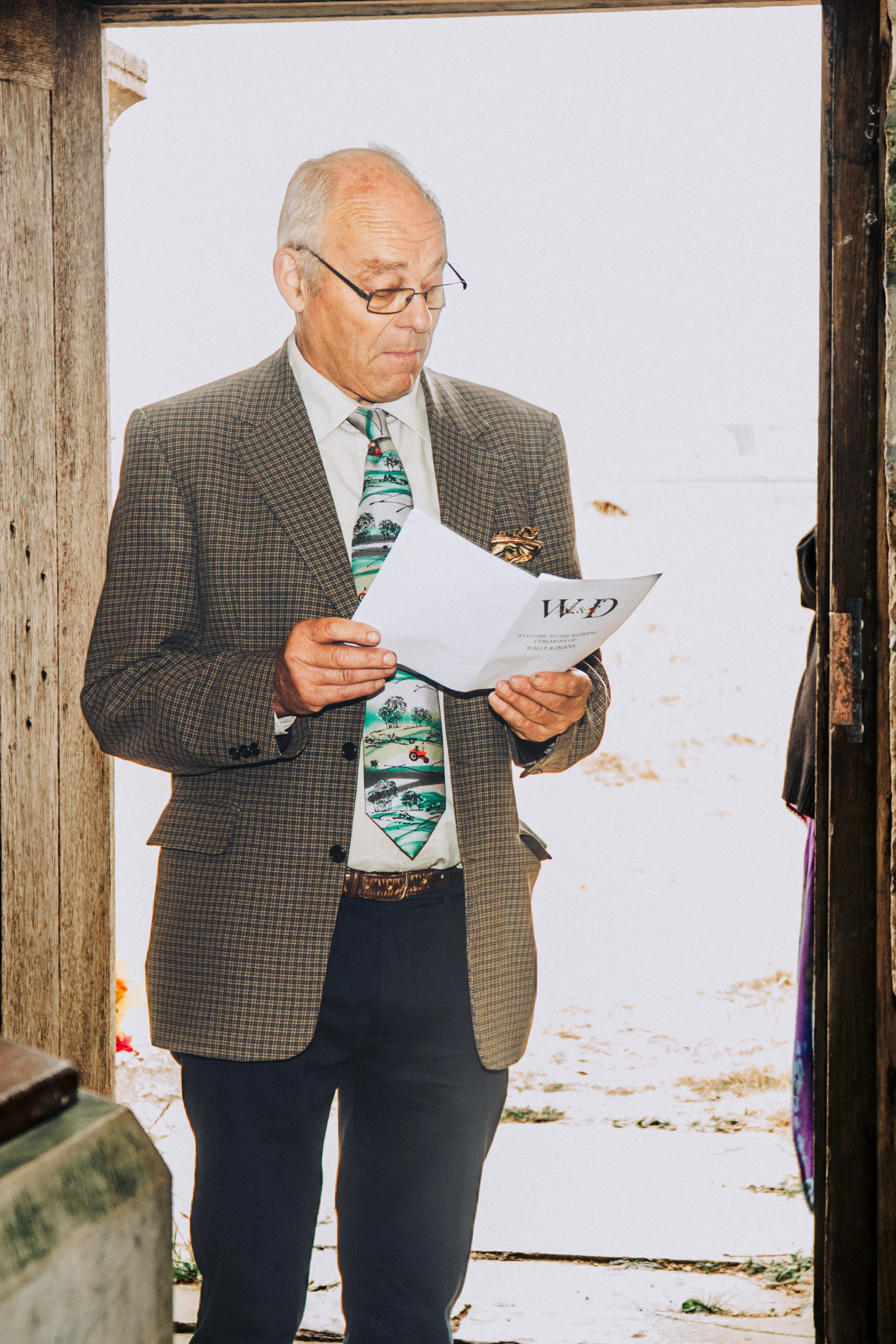 An older male wedding guest wearing a checked suit and patterned tie stands in a doorway, reading through an order of service programme.
