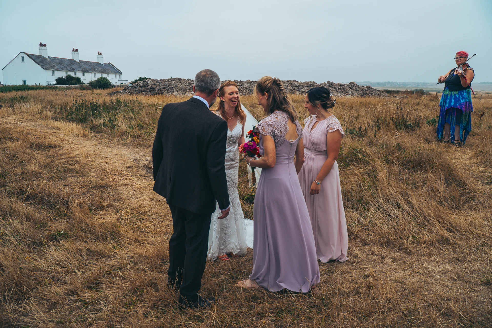 A bride and groom laughing together outdoors in a field alongside bridesmaids and a musician playing the violin under an overcast sky.