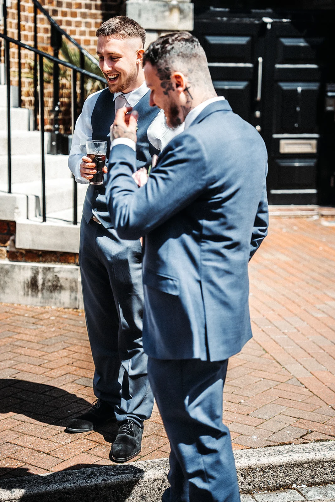 Two groomsmen in blue suits laugh together outside a brick venue while holding drinks on a sunny day.