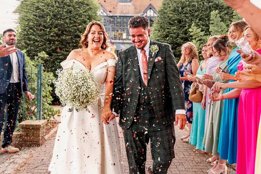 A joyous newlywed couple walks hand in hand through a shower of colourful confetti, surrounded by cheering guests outside a country venue.