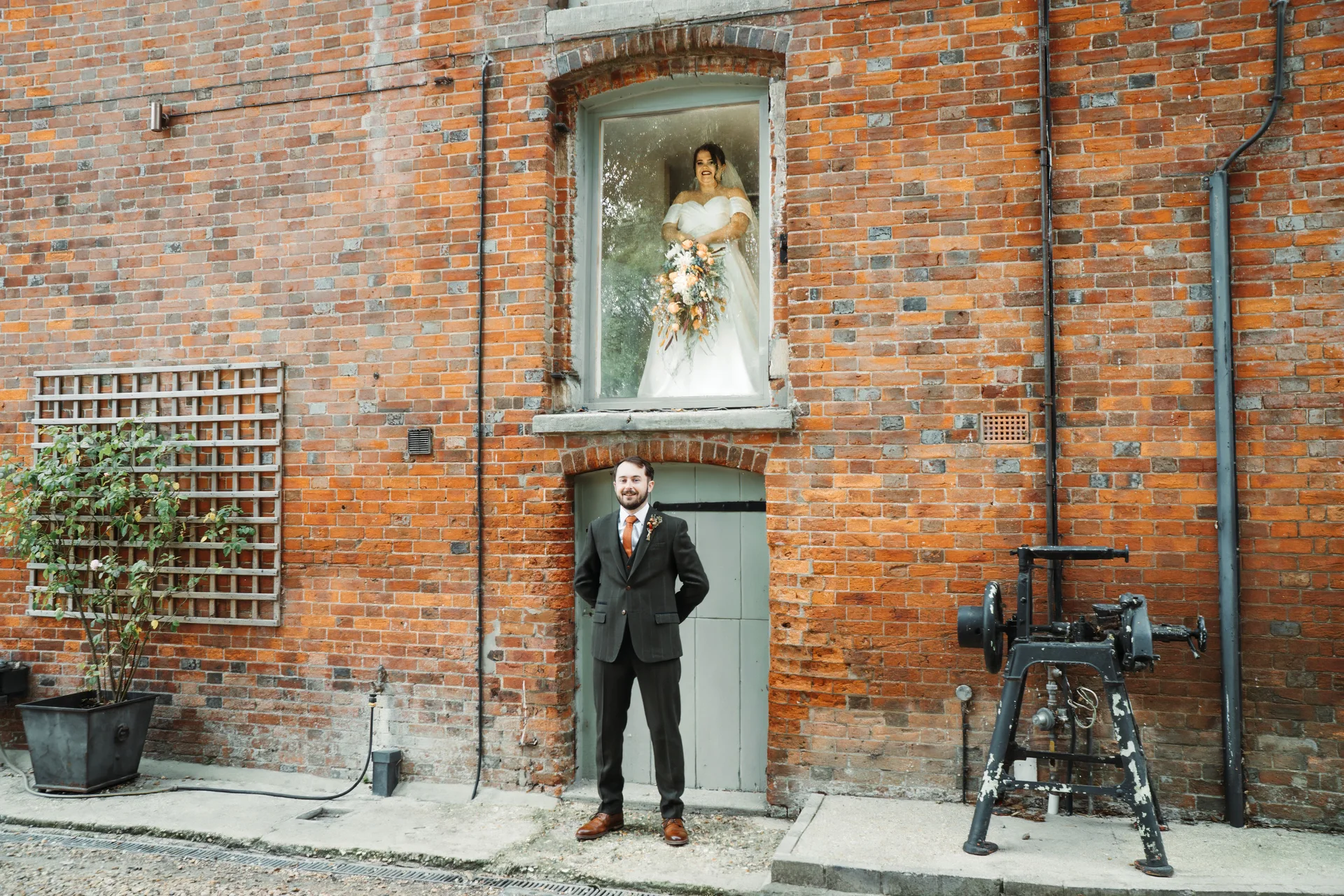 The groom stands outside a rustic brick warehouse doorway while the bride looks down from an upper window holding a cascading floral bouquet.