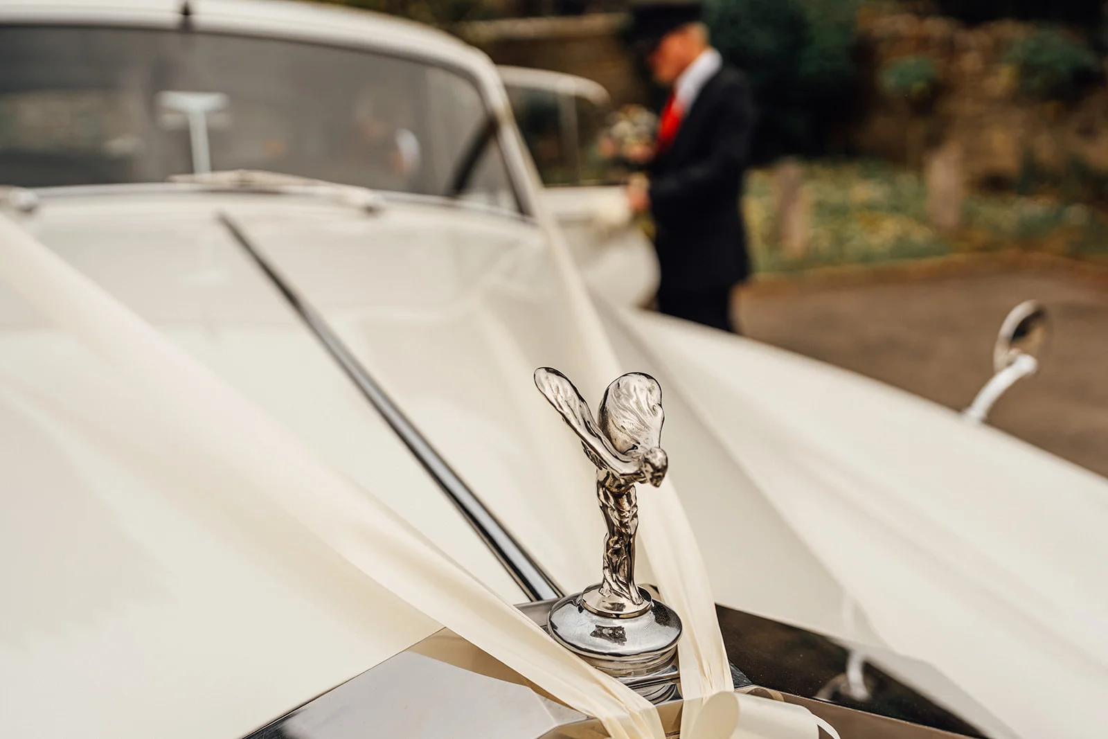 A close-up shot of the silver spirit ornament on the bonnet of a cream wedding car, with a chauffeur standing in the blurred background.