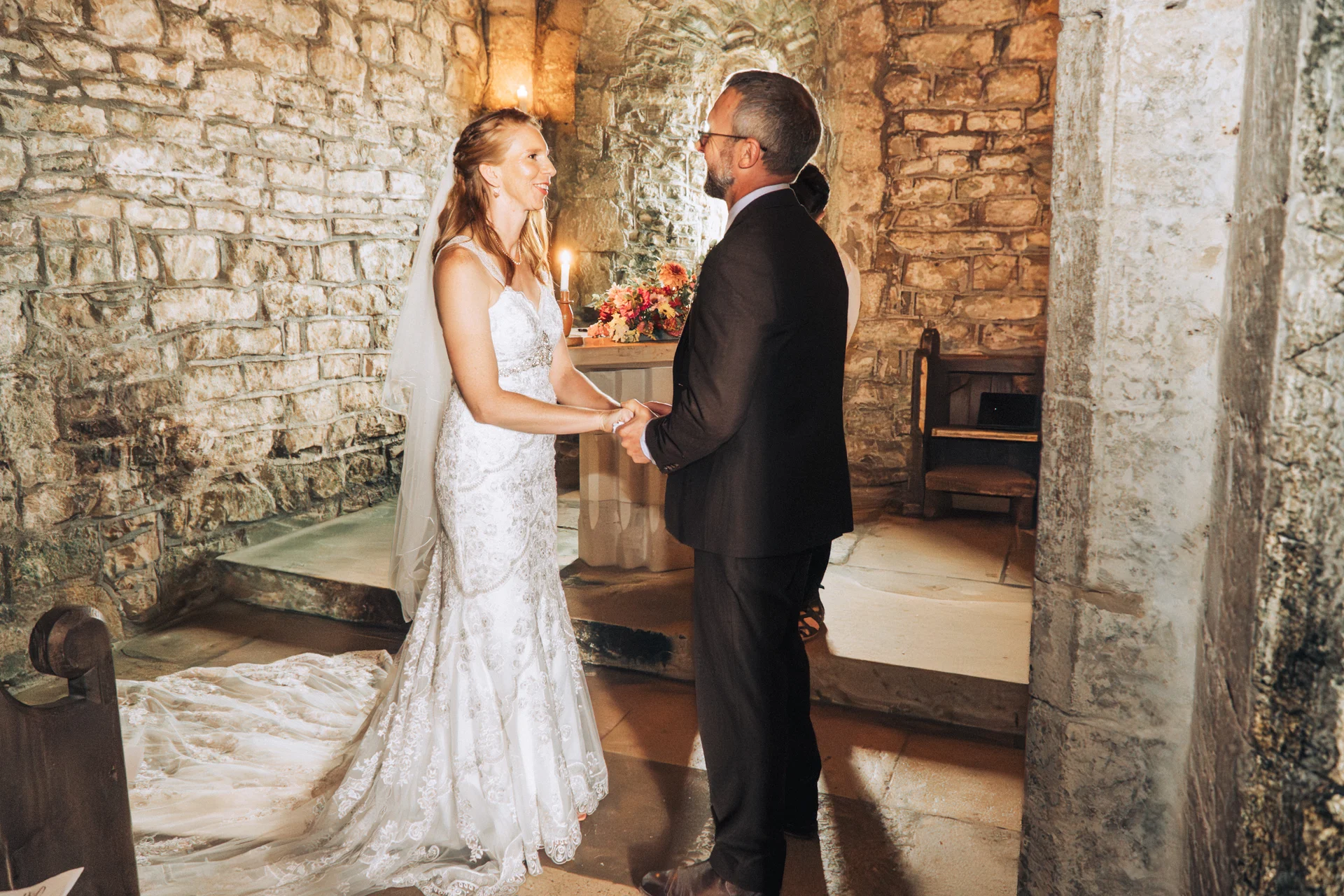 A bride and groom hold hands lovingly during their ceremony inside a historic stone church featuring exposed walls and glowing candlelight.