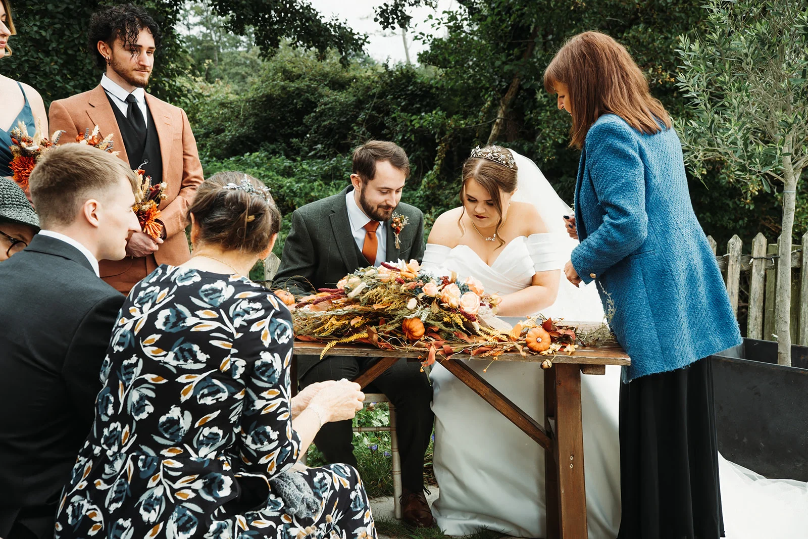 A bride and groom sign the marriage register at an outdoor ceremony table decorated with autumnal foliage and mini pumpkins, surrounded by wedding guests.