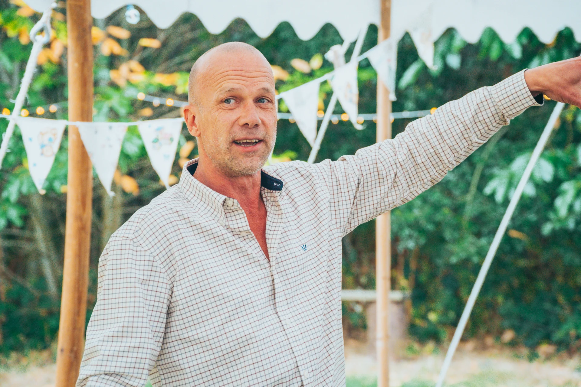 A guest wearing a checked shirt gestures happily inside a marquee decorated with bunting and string lights.