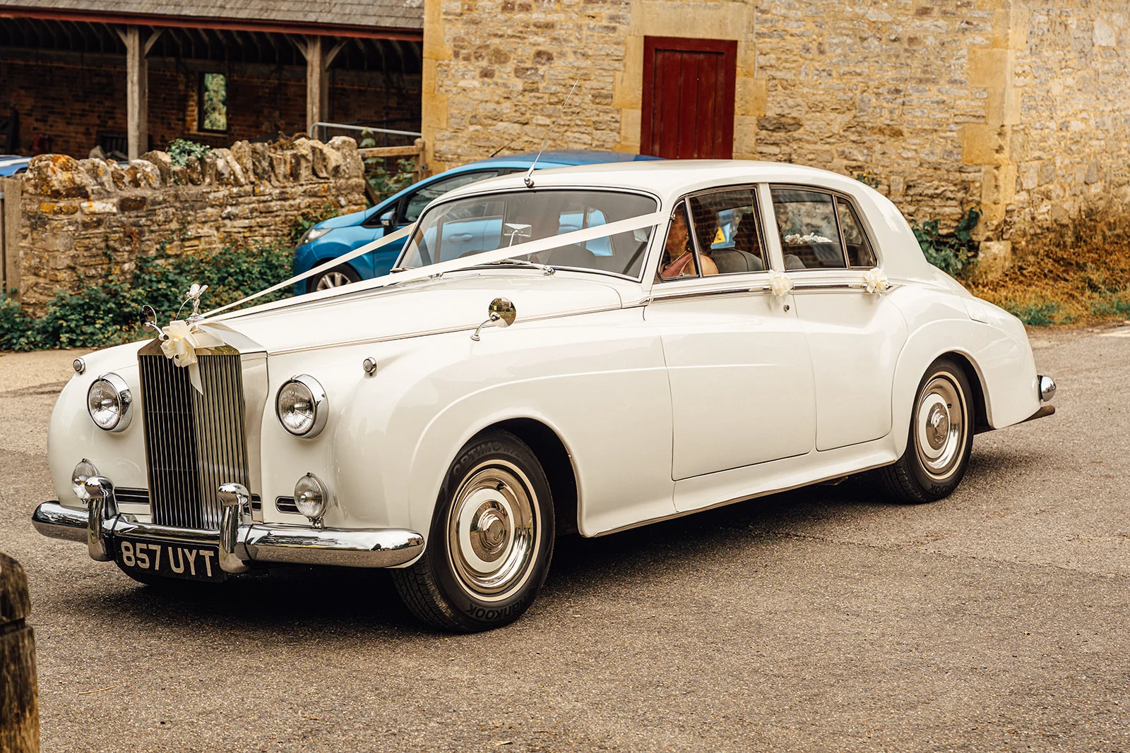 A vintage white wedding car decorated with ribbons is parked outside a rustic stone barn venue.