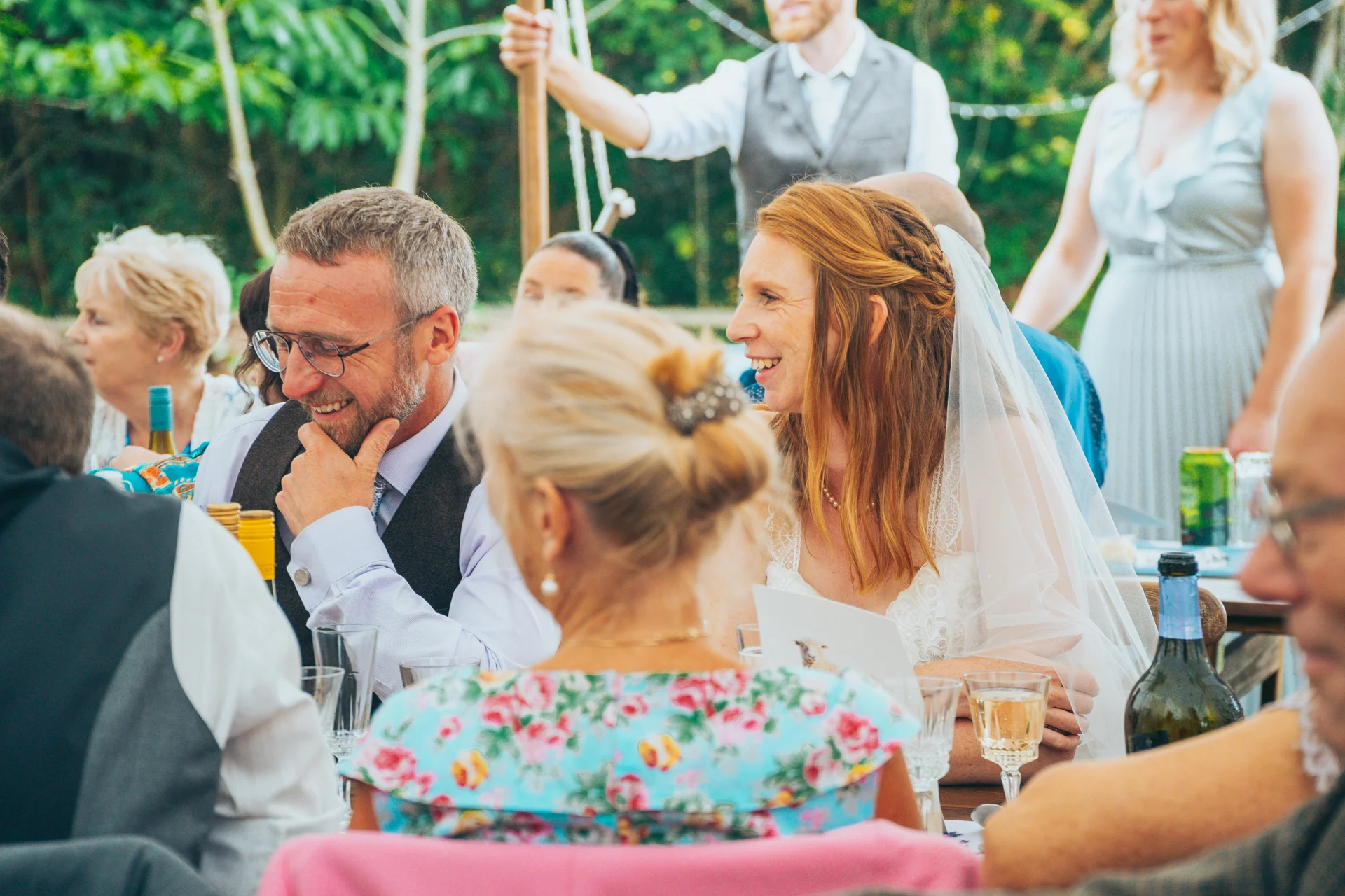 The bride and groom smile and laugh together seated at a banquet table amongst wedding guests during the wedding breakfast.