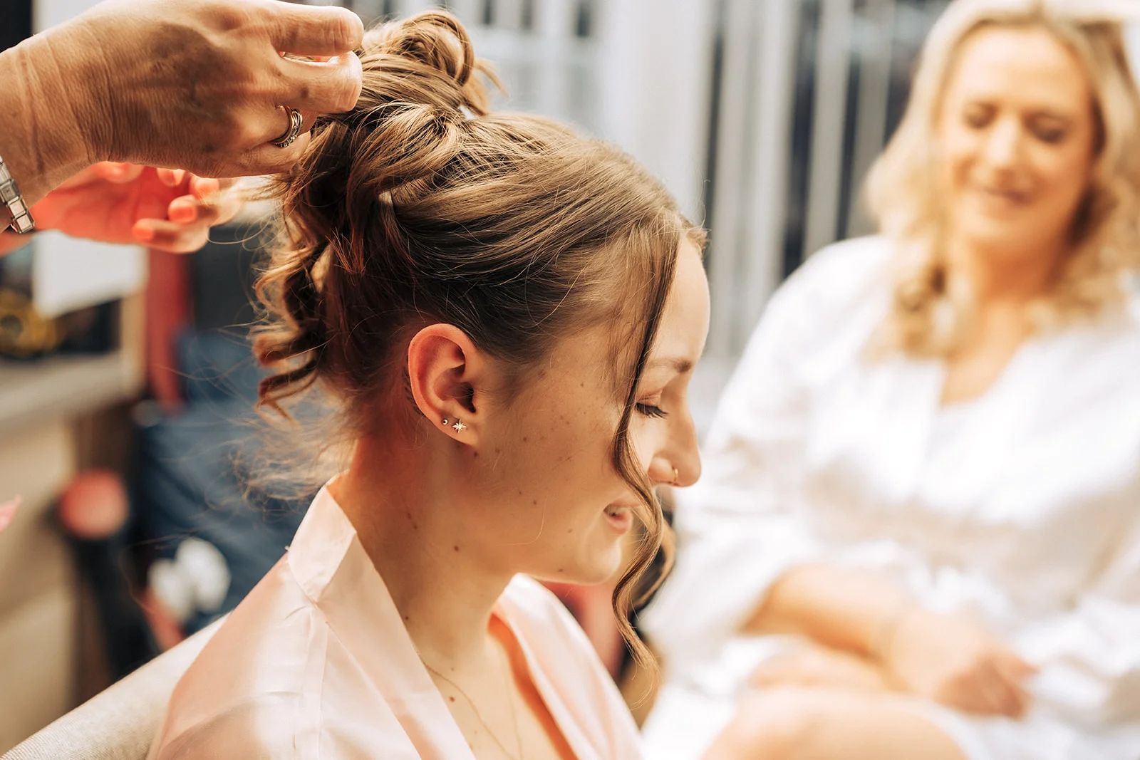 A bride sits calmly as her hairstylist puts the finishing touches to an intricate high bun during morning wedding preparations.