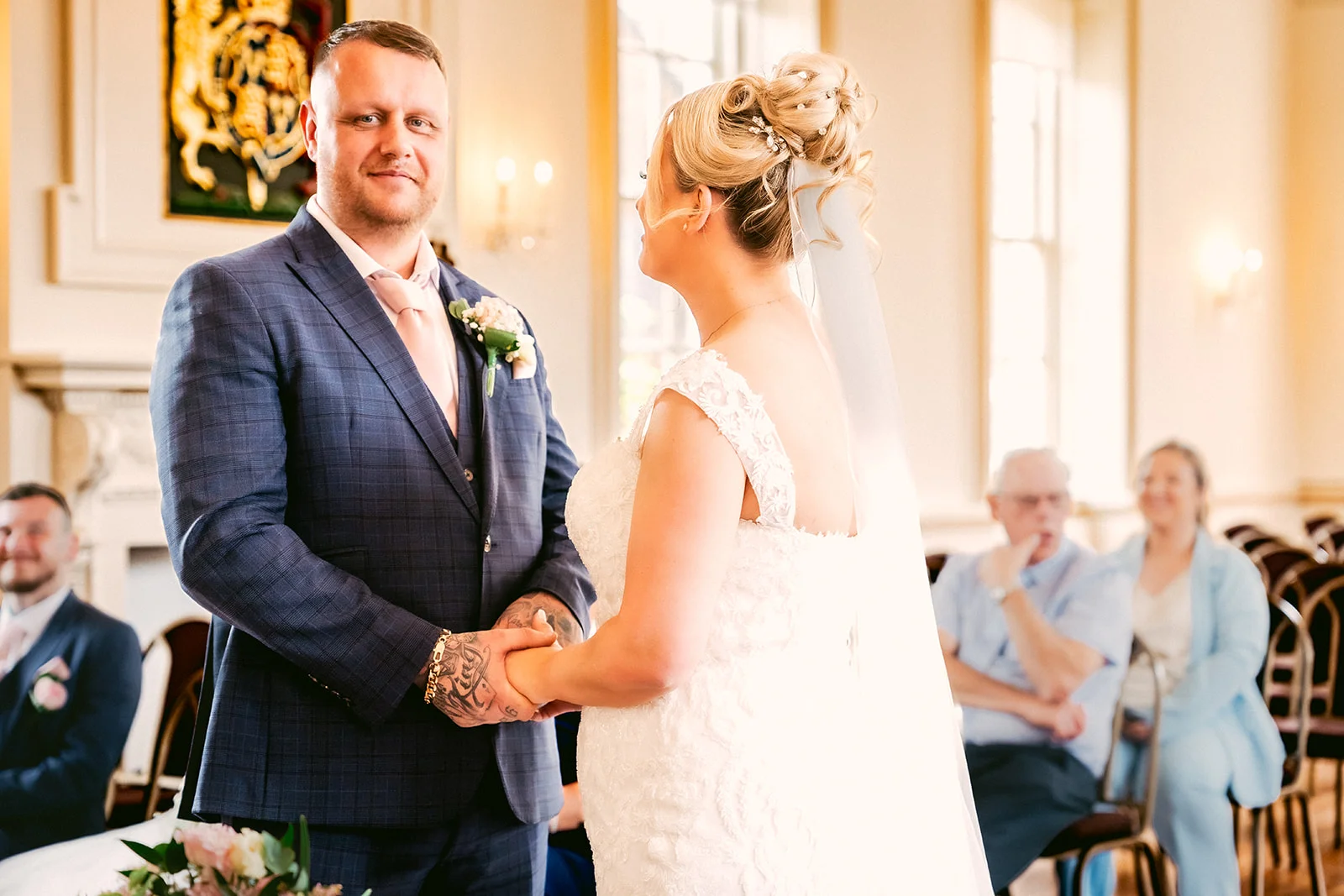 A groom in a blue checked suit and the bride in a lace wedding dress hold hands at the altar during a civil ceremony.