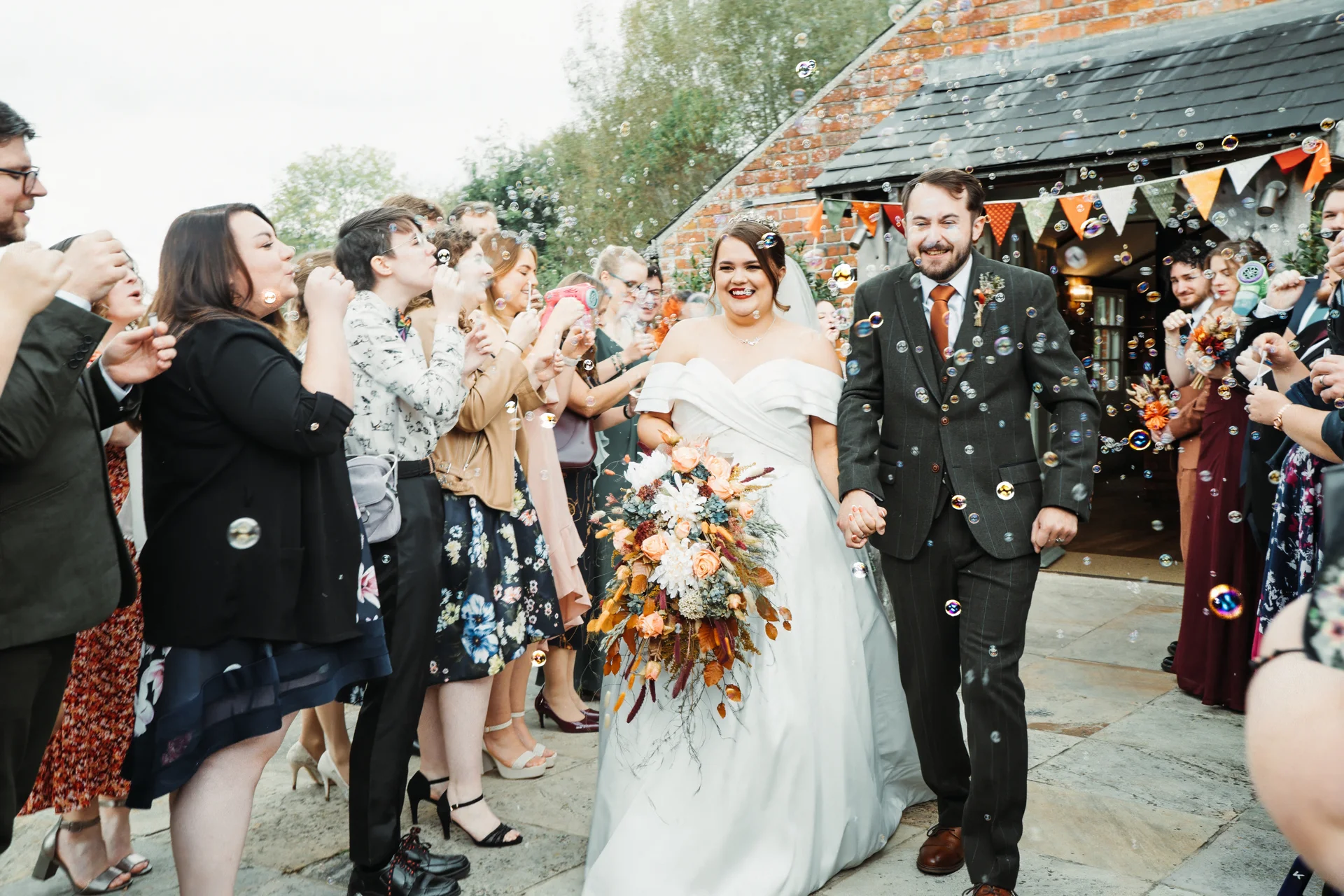 A joyful newlywed couple walks hand in hand past cheering wedding guests blowing bubbles outside a rustic brick barn venue.