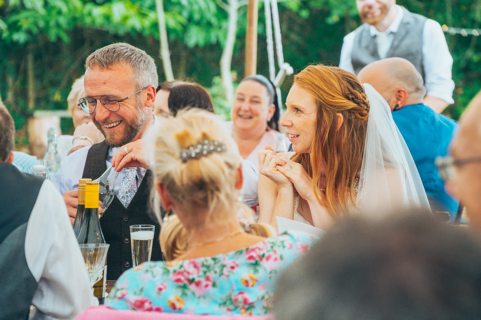 The bride and groom laugh together at the wedding breakfast table surrounded by happy guests enjoying speeches outdoors.