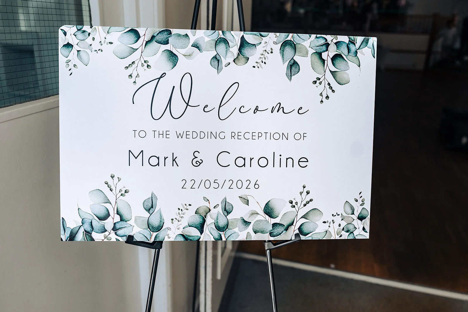 An elegant wedding welcome sign featuring modern calligraphy and eucalyptus leaf illustrations, resting on a black metal easel.