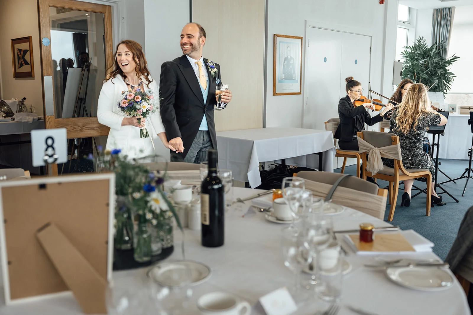 A smiling bride and groom enter a reception room holding hands, walking past decorated dining tables while a string quartet plays in the background.