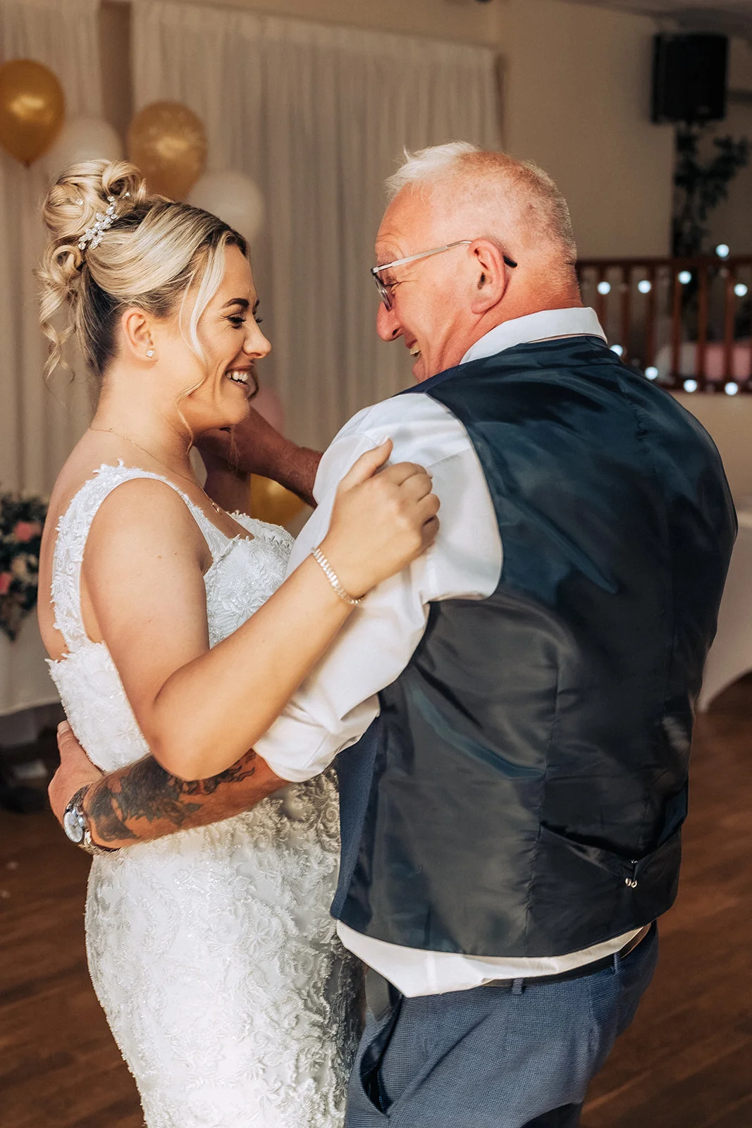 A smiling bride shares a joyful father daughter dance with her father, who wears a waistcoat and glasses during the wedding reception.
