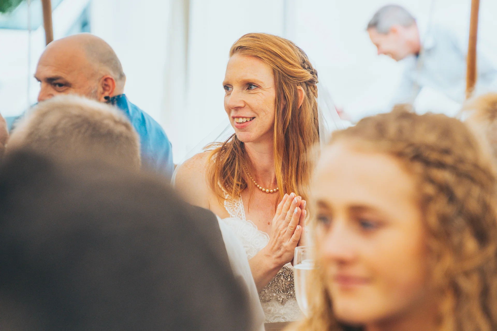 A smiling red-haired bride with freckles and a pearl necklace claps happily amongst wedding guests during the reception speeches.