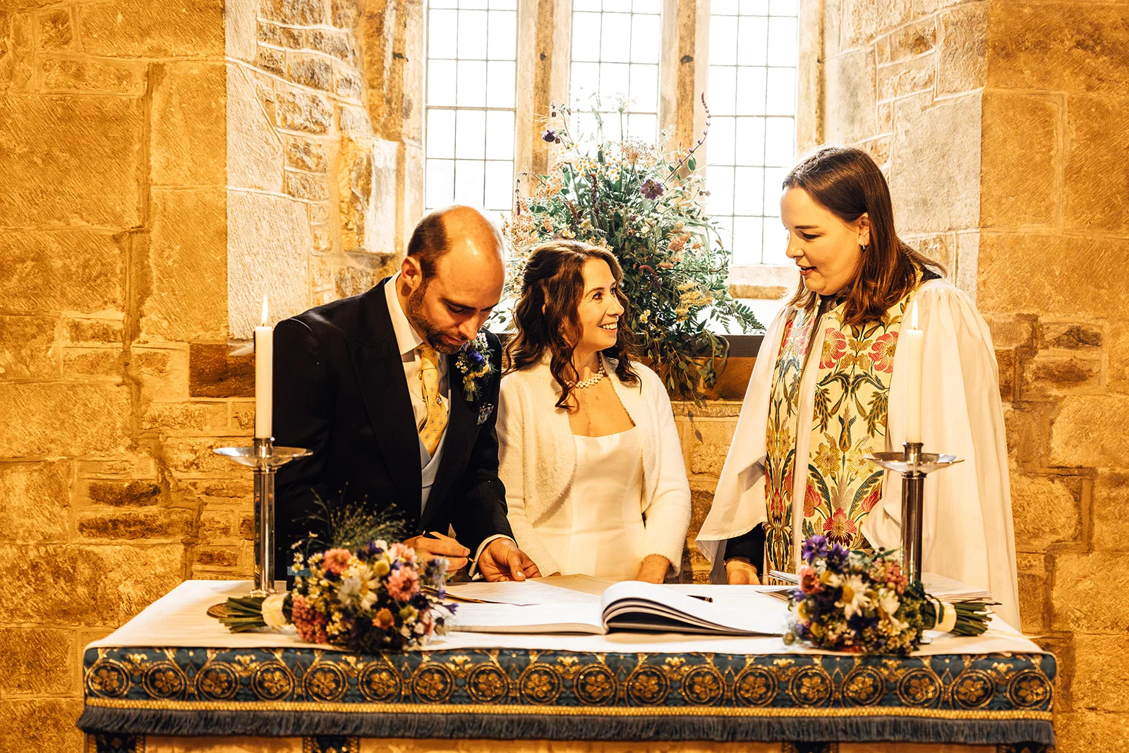 The bride and groom sign the marriage register at the altar inside a historic stone church, accompanied by the officiant.
