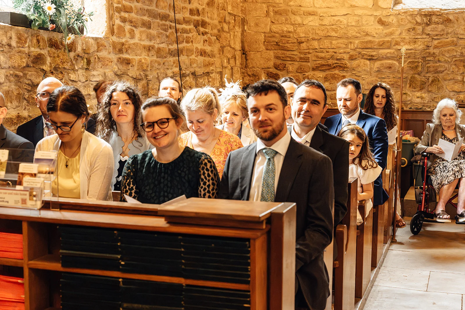 A congregation of smiling wedding guests sits in wooden pews inside a historic stone church during the ceremony.