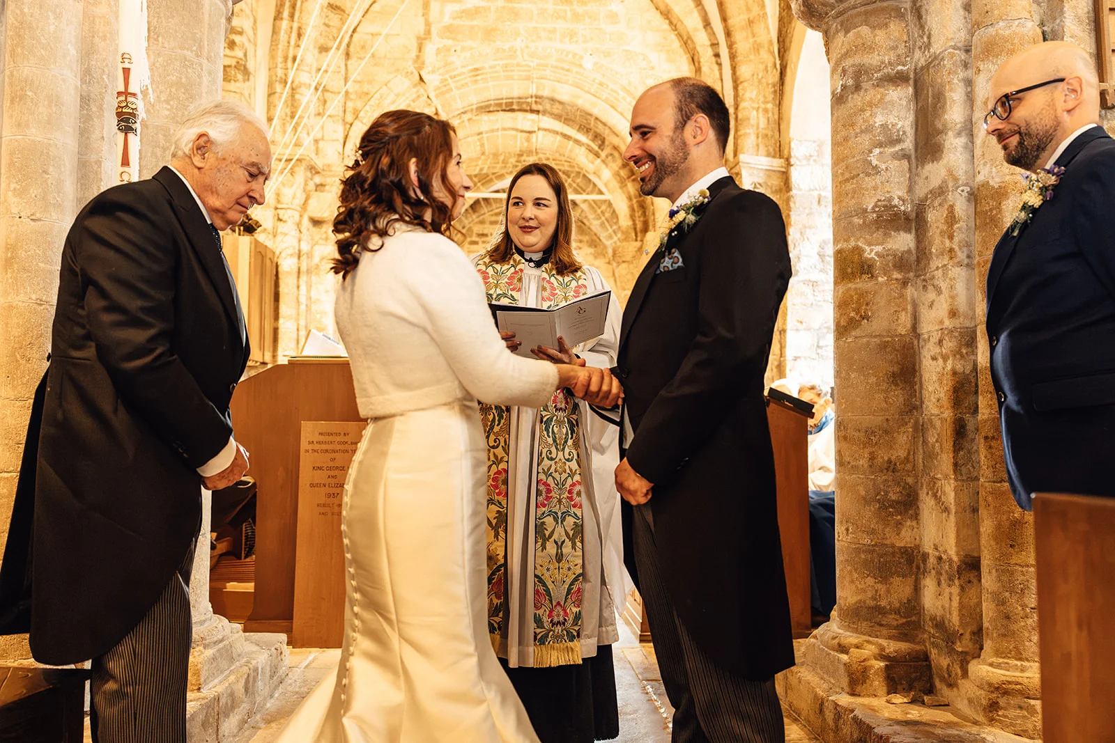 A bride and groom lovingly hold hands during their church ceremony, flanked by family members and the celebrant inside a historic stone building.