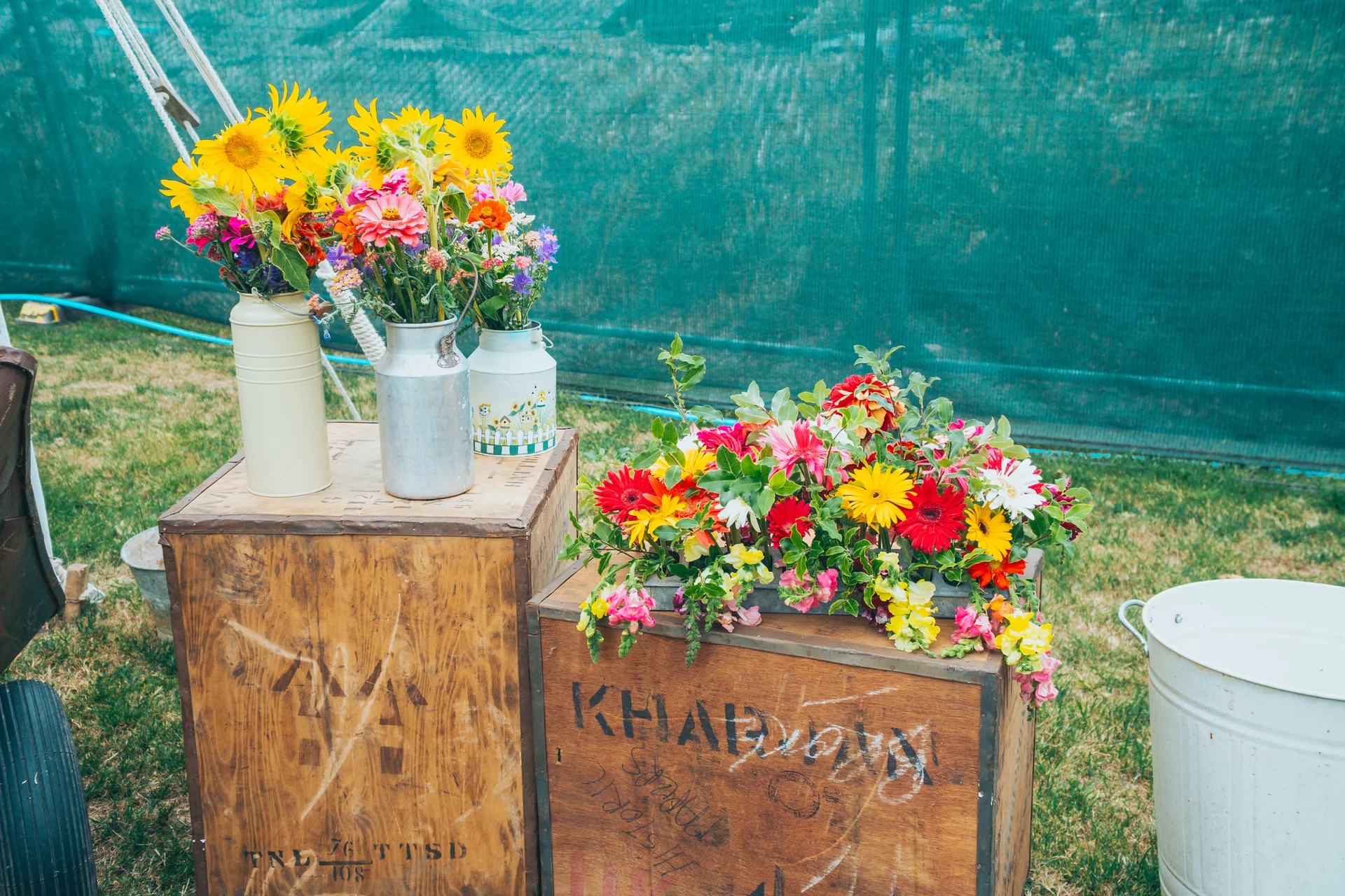 A vibrant rustic display of colourful wildflowers and sunflowers arranged in milk churns and wooden crates against a green fabric backdrop.