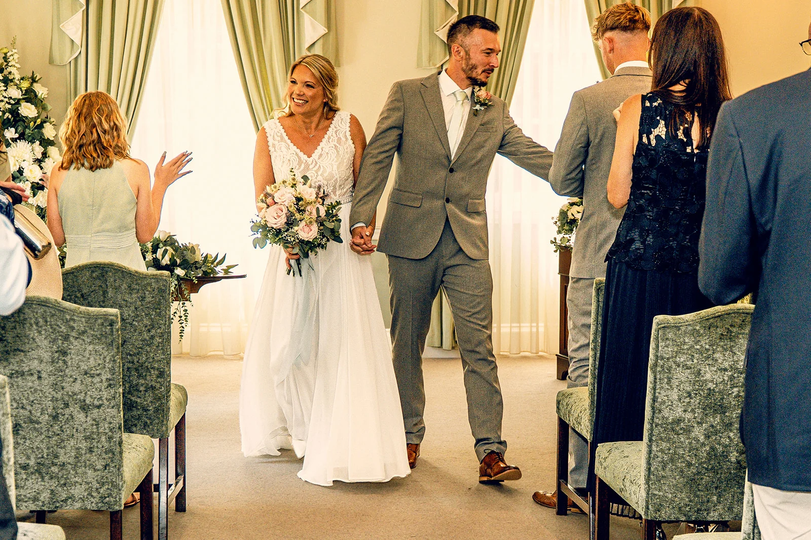 Newlywed bride and groom walk hand in hand down the aisle smiling while guests applaud.