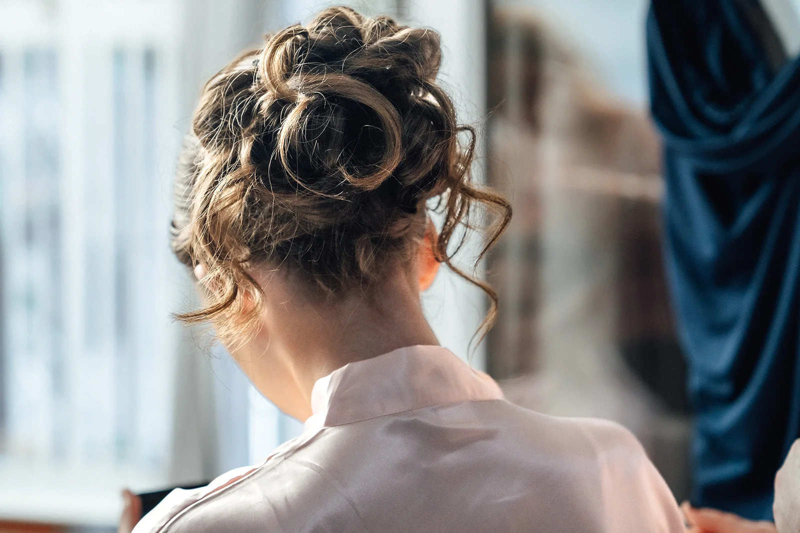A close-up back view of a bride with a textured updo hairstyle and face-framing tendrils, wearing a pale pink satin robe during morning preparations.