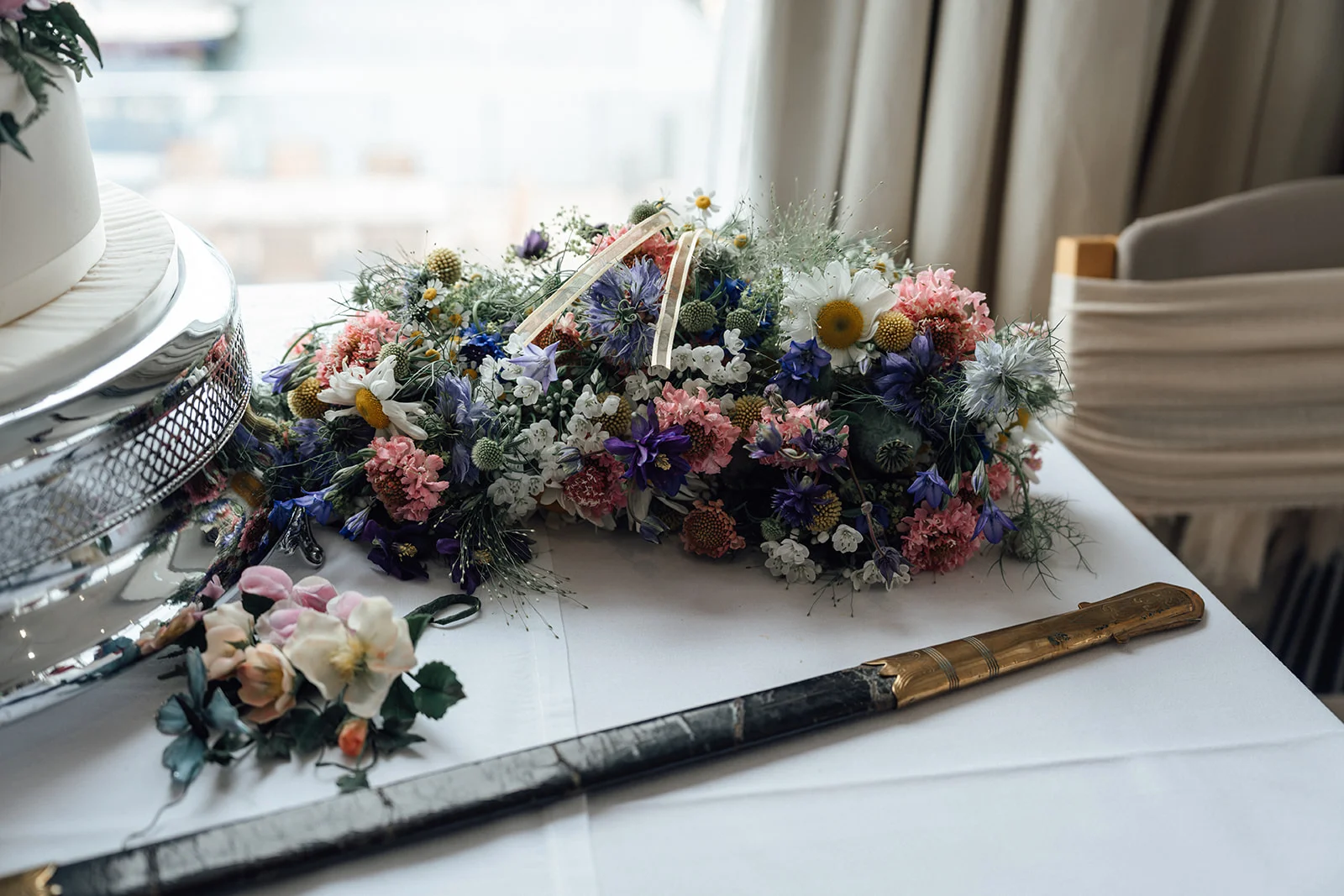 A detailed close-up shot shows a wild unstructured wildflower bouquet resting on a white linen-covered table next to a ceremonial sword scabbard and a silver cake stand.