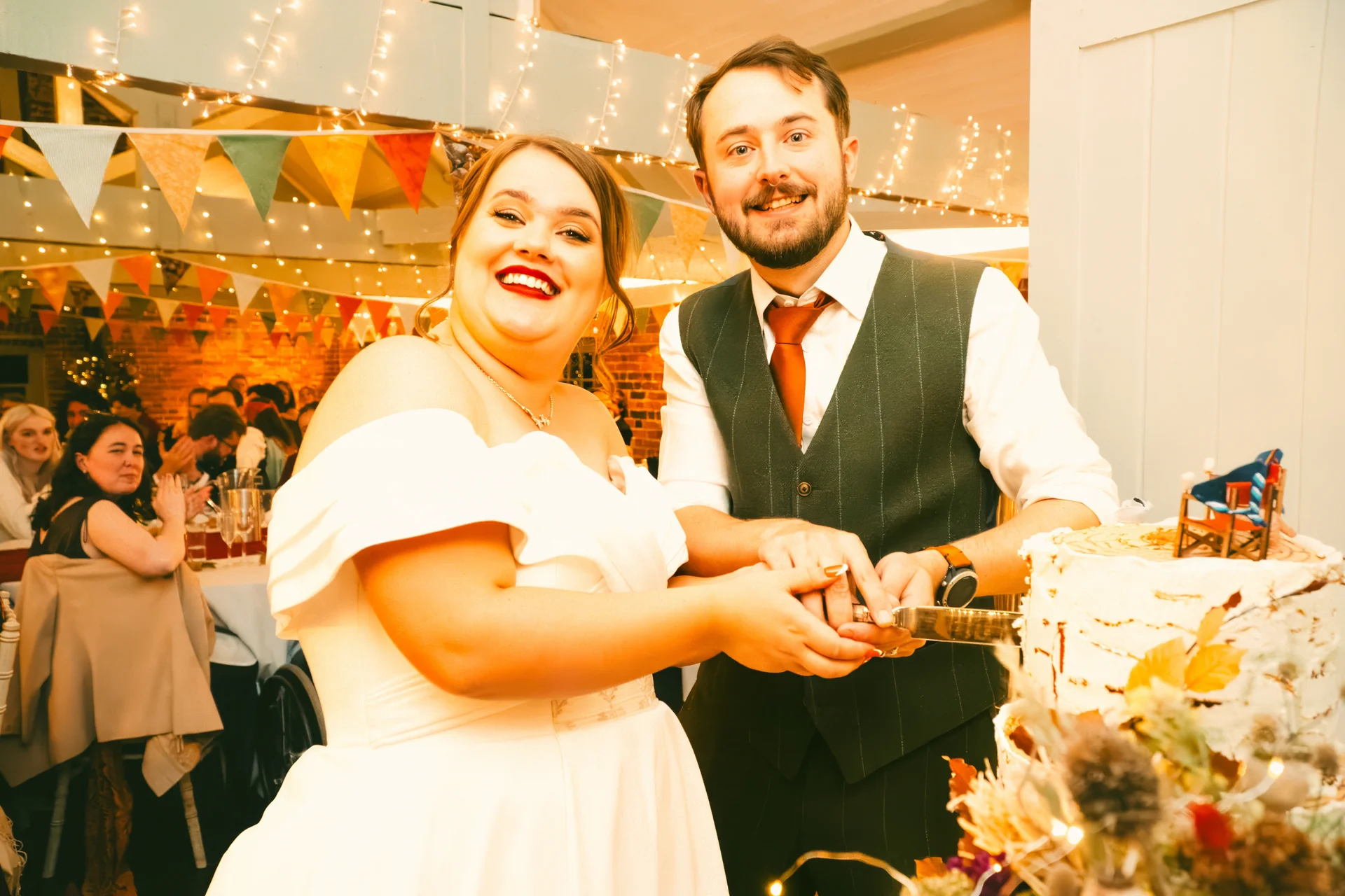 A newlywed couple happily cut their wedding cake together in a rustic barn venue decorated with colourful bunting and warm fairy lights.