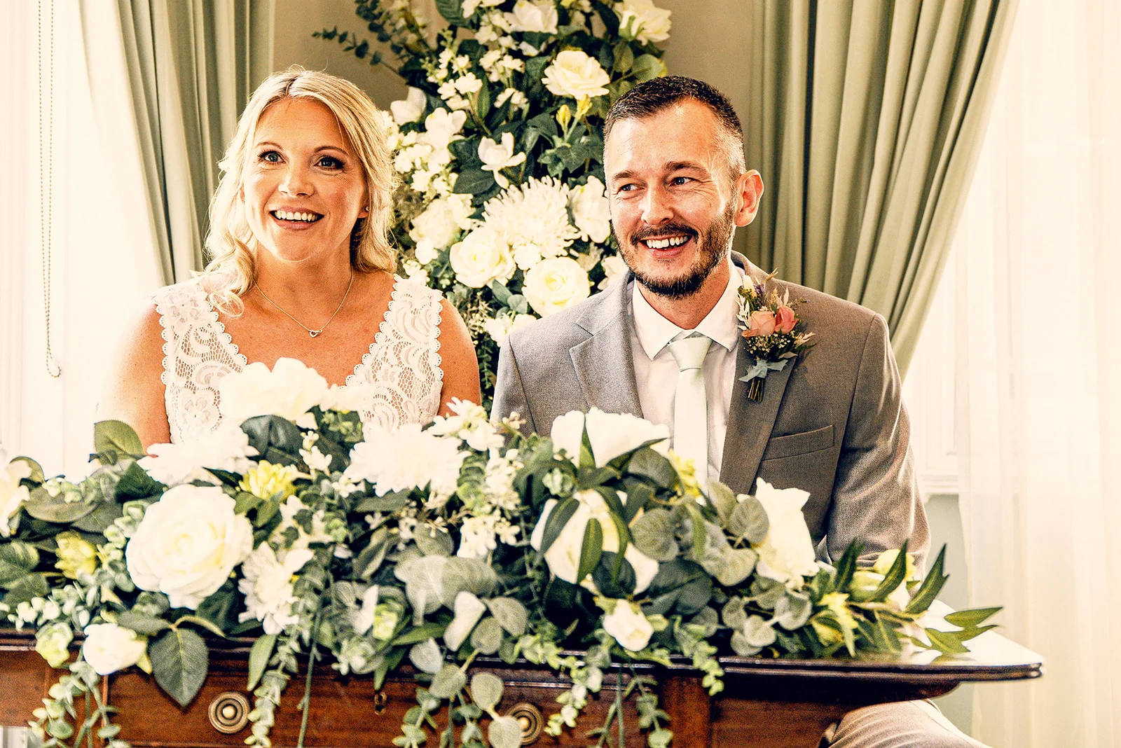 A smiling bride and groom sit together behind a floral-decorated table during their wedding ceremony, framed by white roses and greenery.