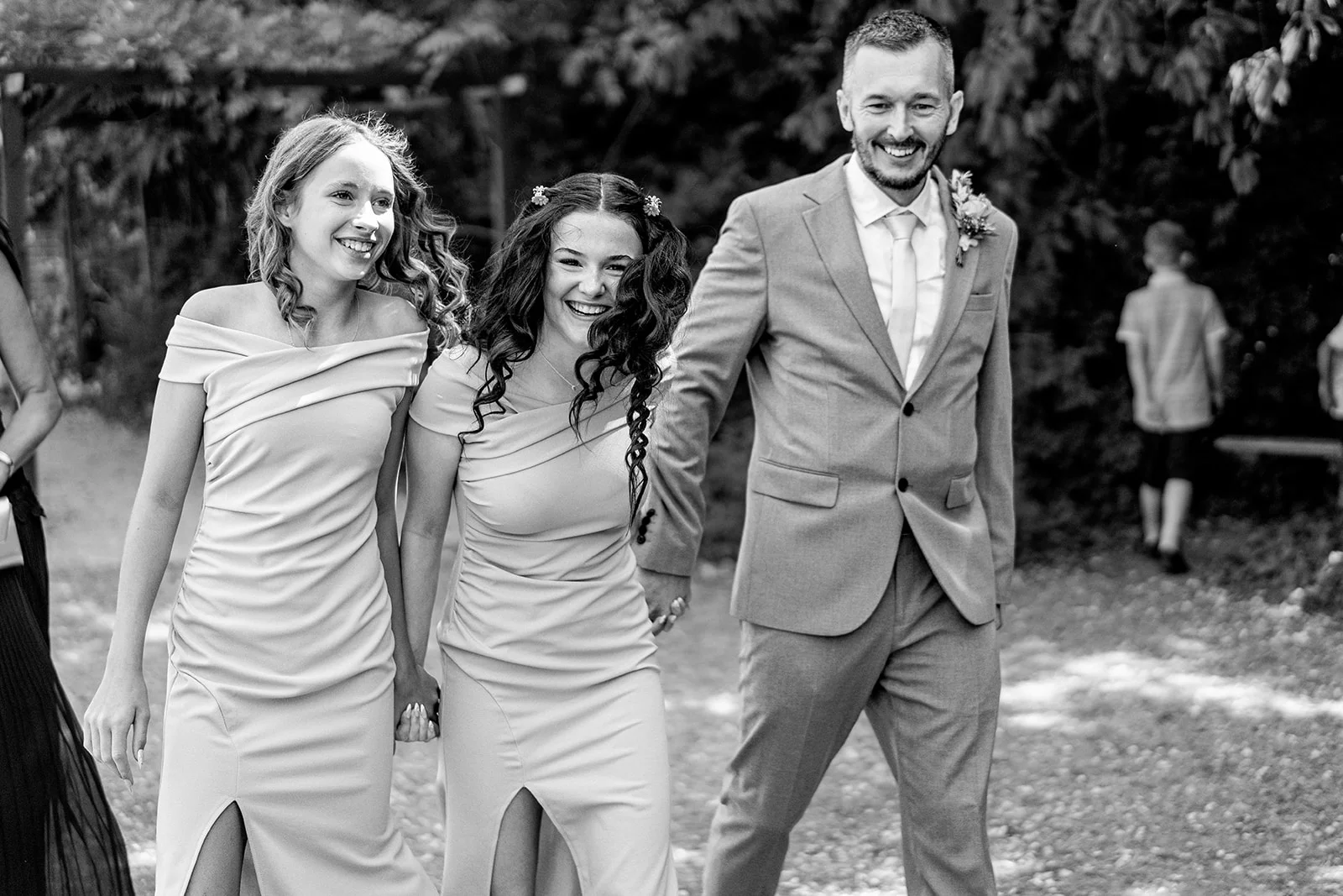 A smiling groom in a grey suit walks hand in hand with two bridesmaids wearing matching off-the-shoulder dresses with thigh splits outdoors.