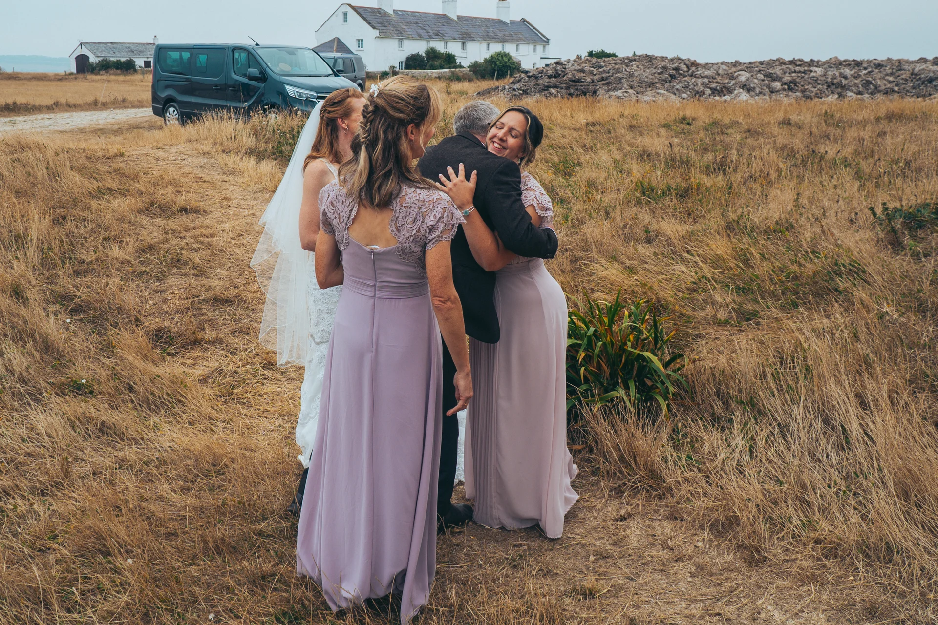 A bride in a white gown and veil shares a joyful embrace with family members outdoors in a rustic meadow setting.