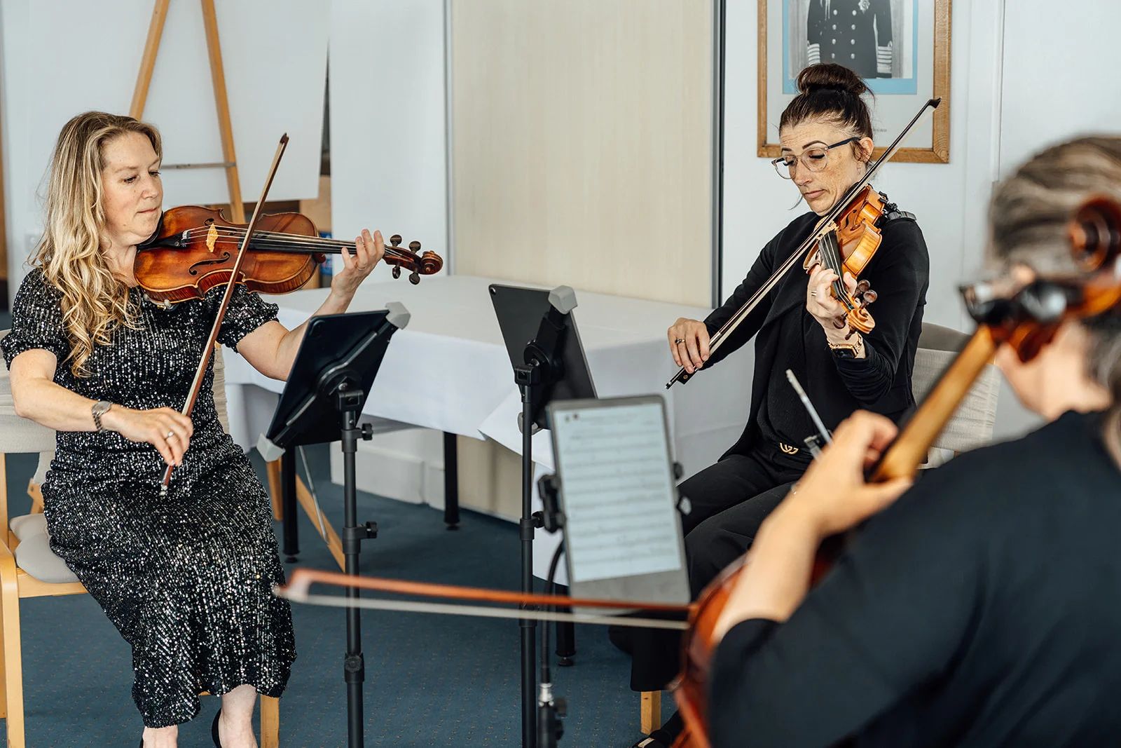 A string trio of female musicians play their instruments during a wedding ceremony in a bright indoor venue.