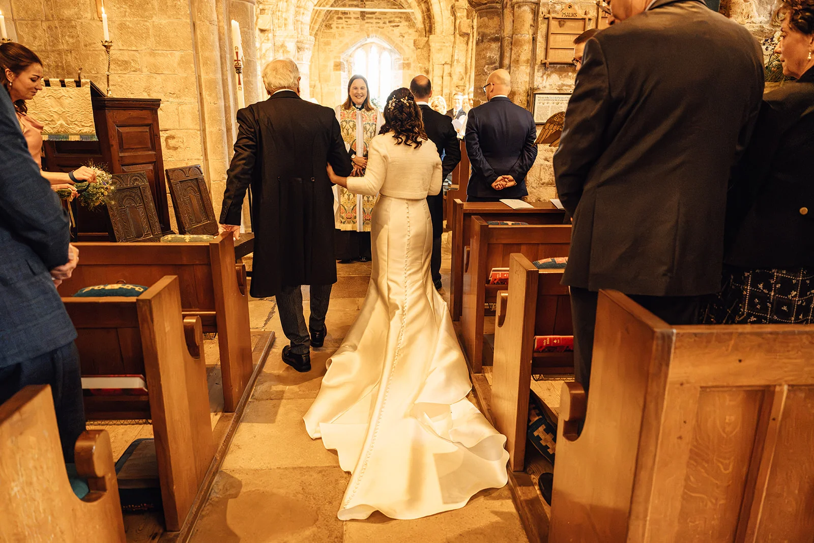 A bride in a satin gown with a long train walks down the aisle of a historic stone church escorted by her father.