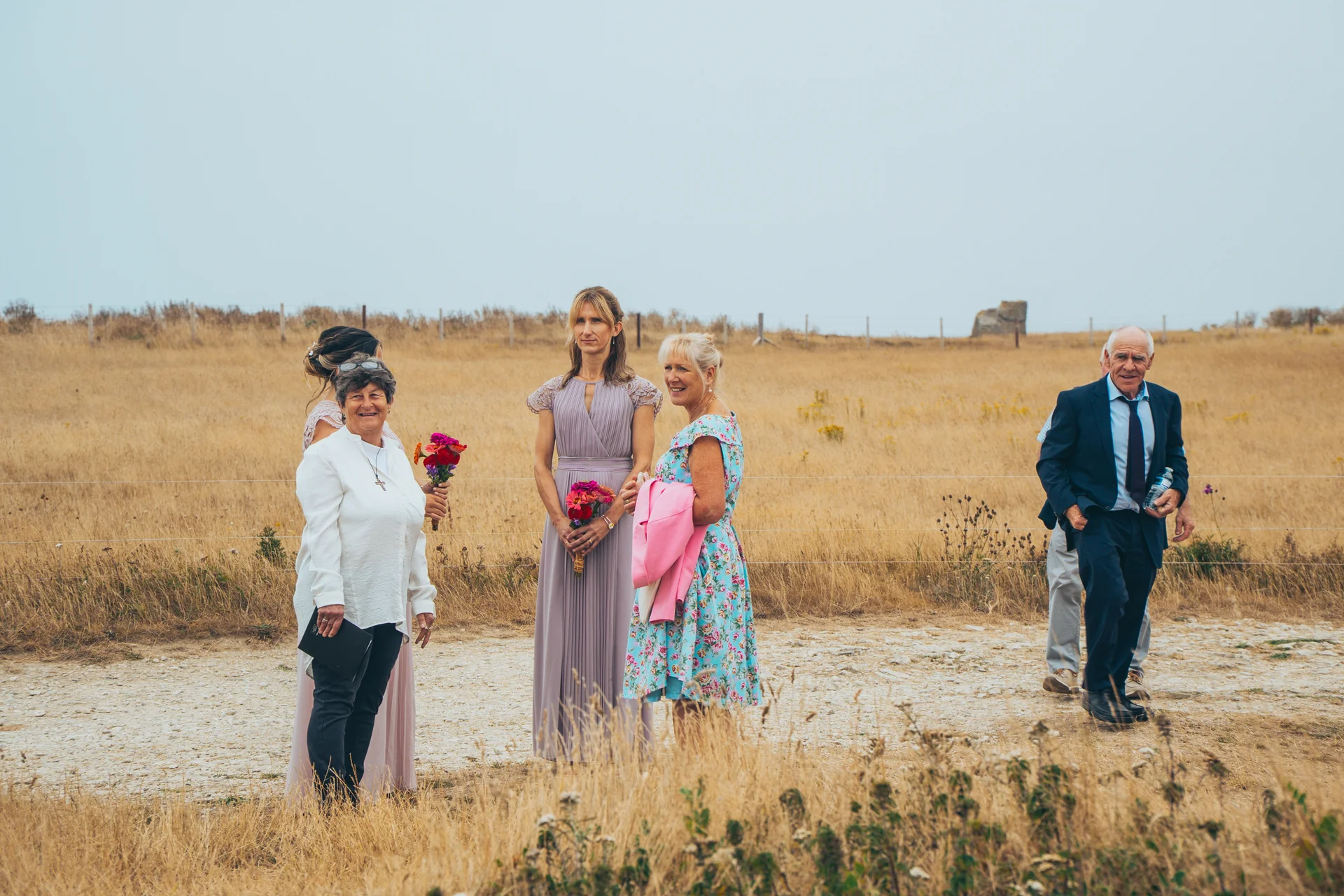 Wedding guests in colourful outfits and suits gather along a coastal path overlooking dry grassy fields under an overcast sky.