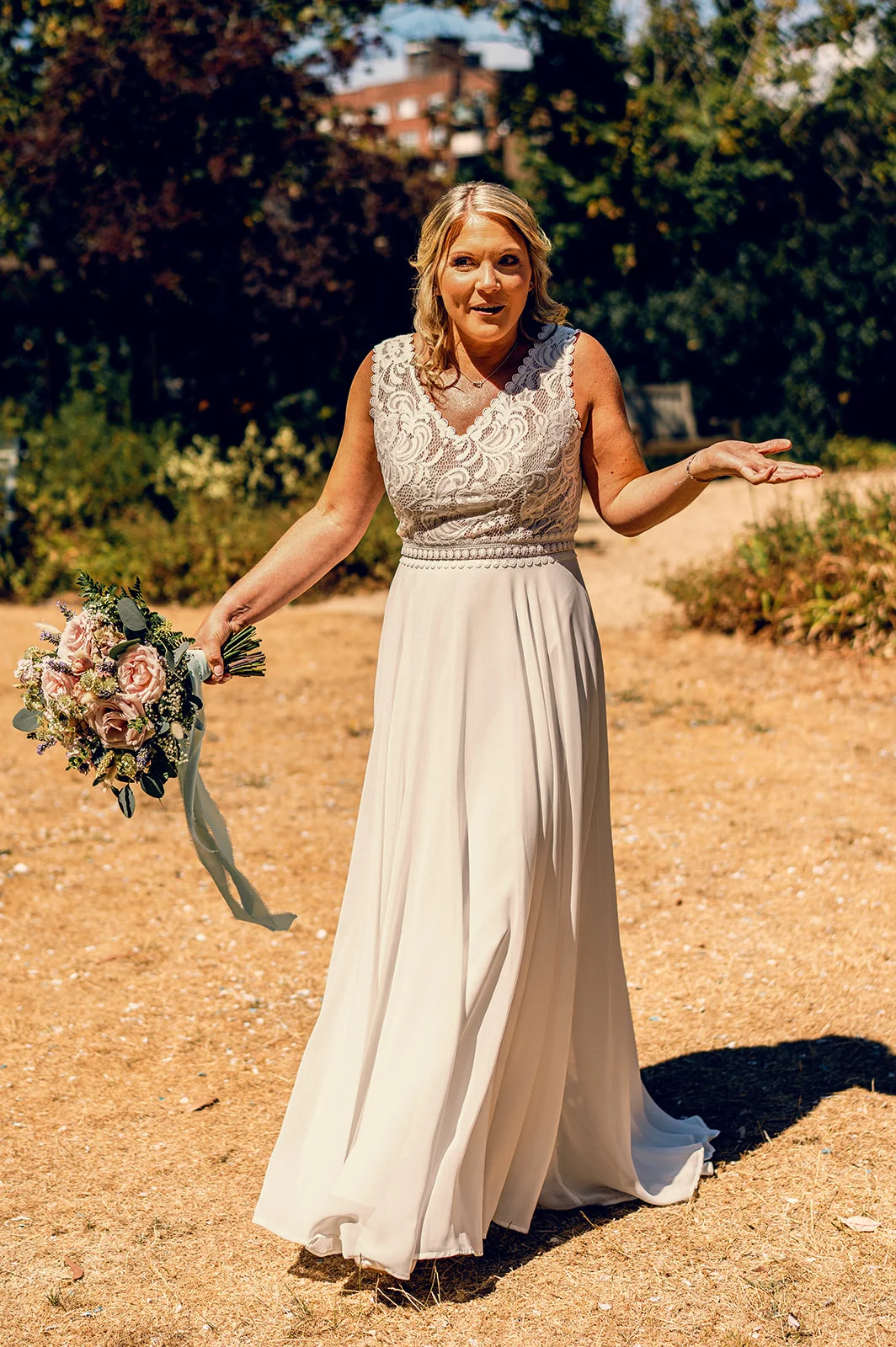 A joyful bride in a lace and chiffon wedding dress laughs outdoors in the sunshine, holding a hand-tied pastel bouquet.