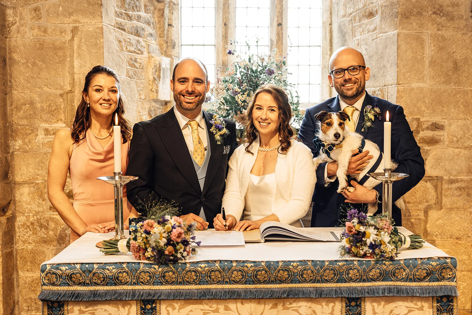A smiling couple signs the wedding register in a historic stone church, accompanied by family, a small Jack Russell terrier in a bow tie, and delicate floral arrangements.