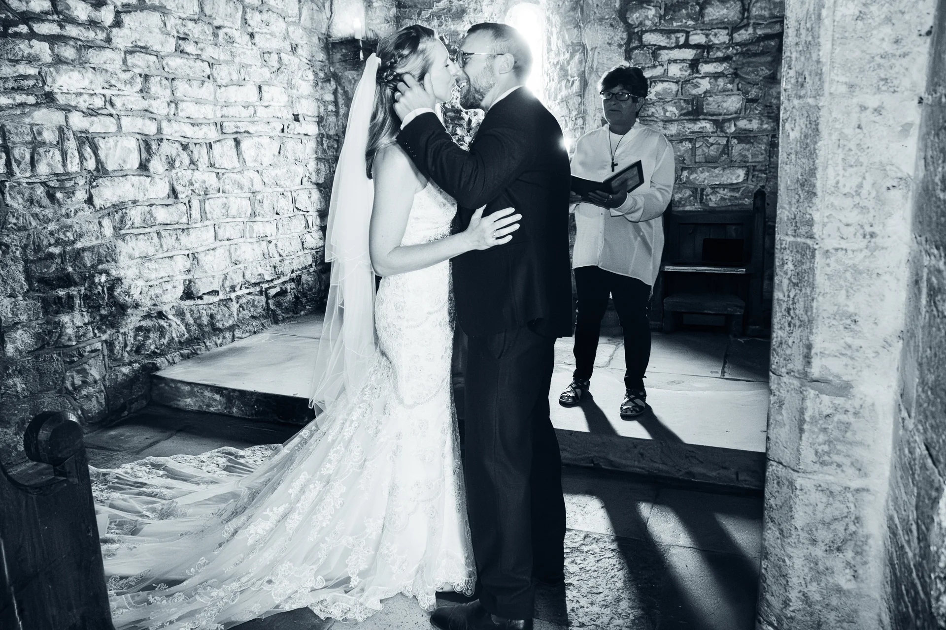 A newlywed couple share their first kiss during a ceremony inside a rustic stone church, captured in a striking black and white photograph.
