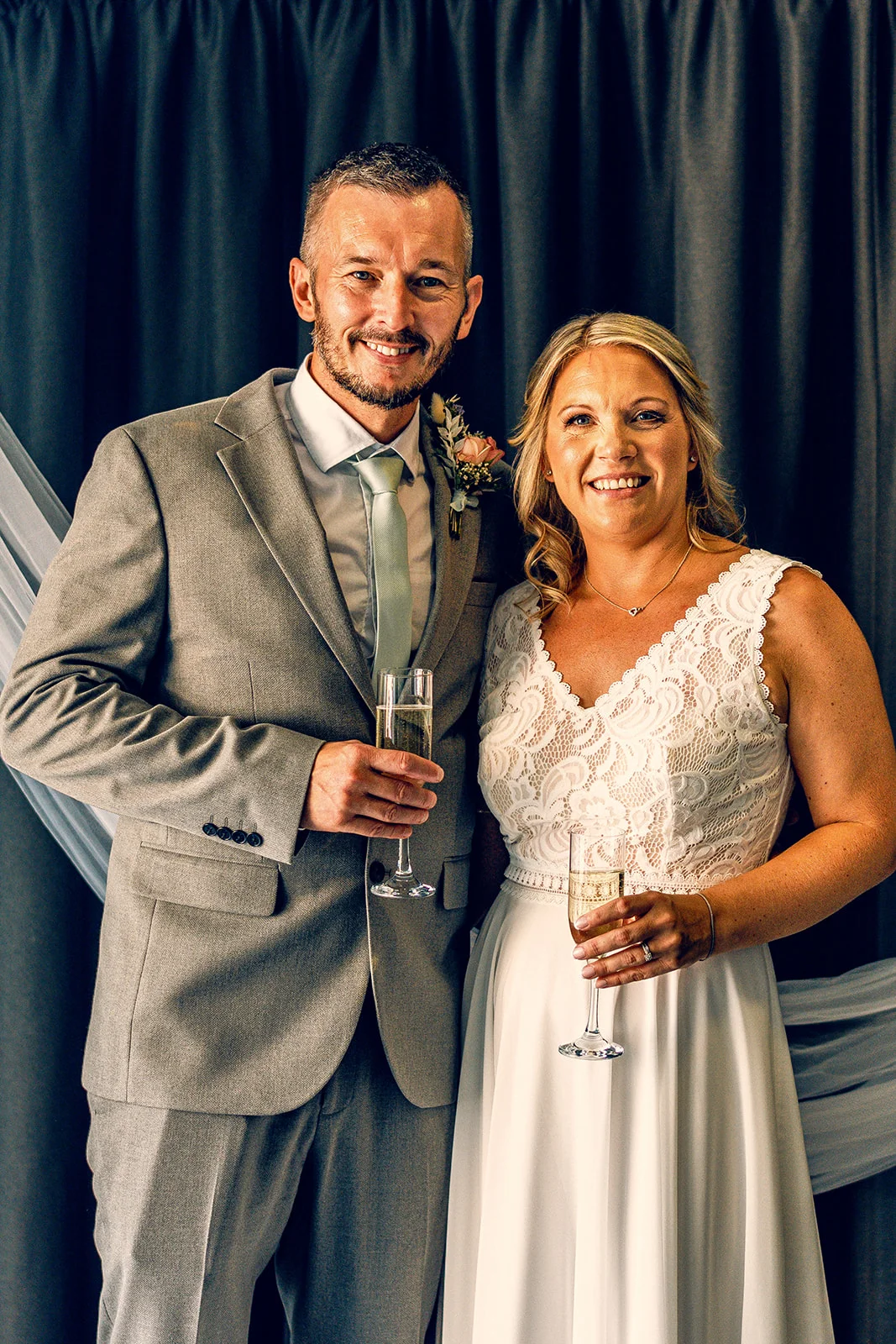 A smiling bride in a lace top wedding dress and a groom in a grey suit pose together holding champagne flutes indoors against a dark curtain backdrop.