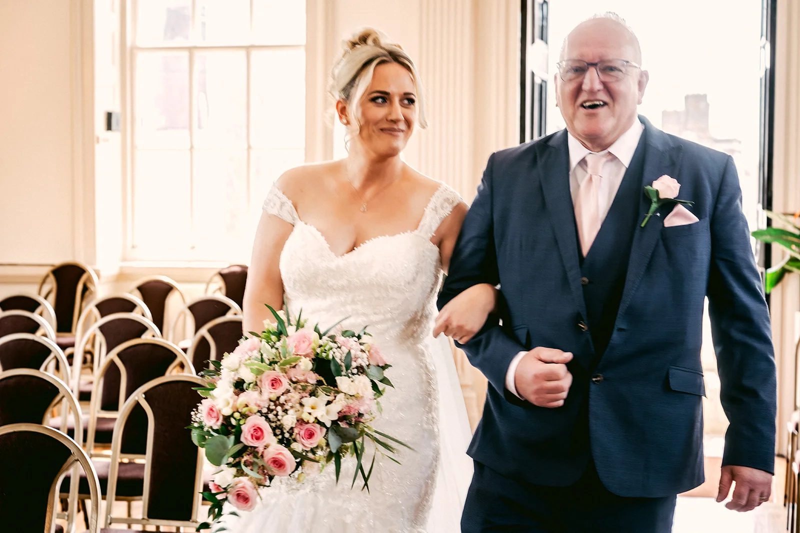 A smiling bride walks down the aisle arm in arm with her father, carrying a cascading pink rose bouquet in a light-filled venue.