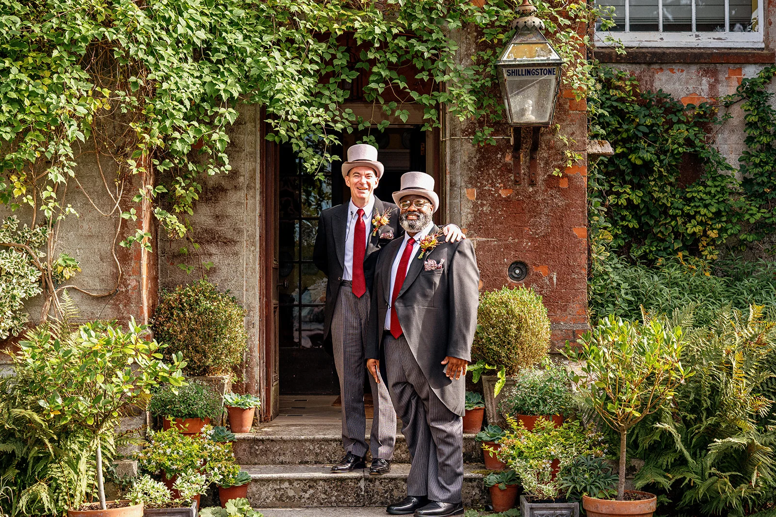 Two grooms dressed in traditional morning suits and top hats pose warmly together in front of an ivy-covered doorway at a country house venue.