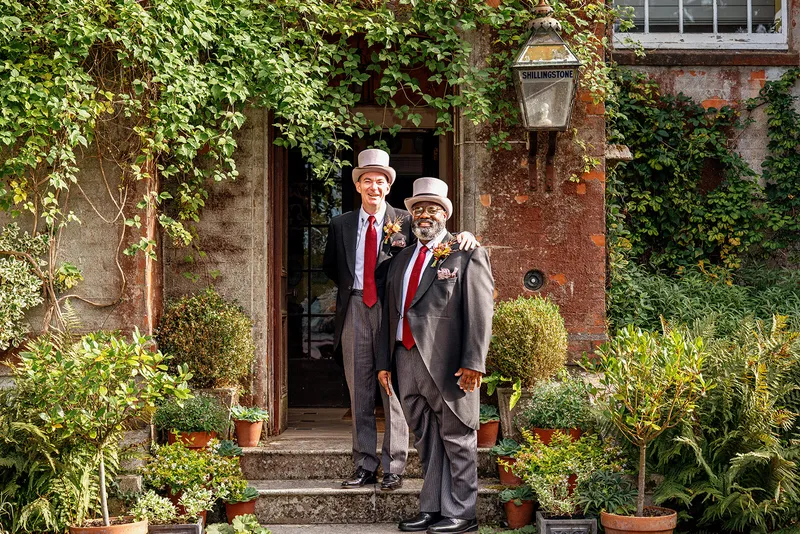 Two grooms dressed in traditional morning suits and top hats pose warmly together in front of an ivy-covered doorway at a country house venue.