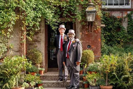 Two grooms dressed in traditional morning suits and top hats pose warmly together in front of an ivy-covered doorway at a country house venue.