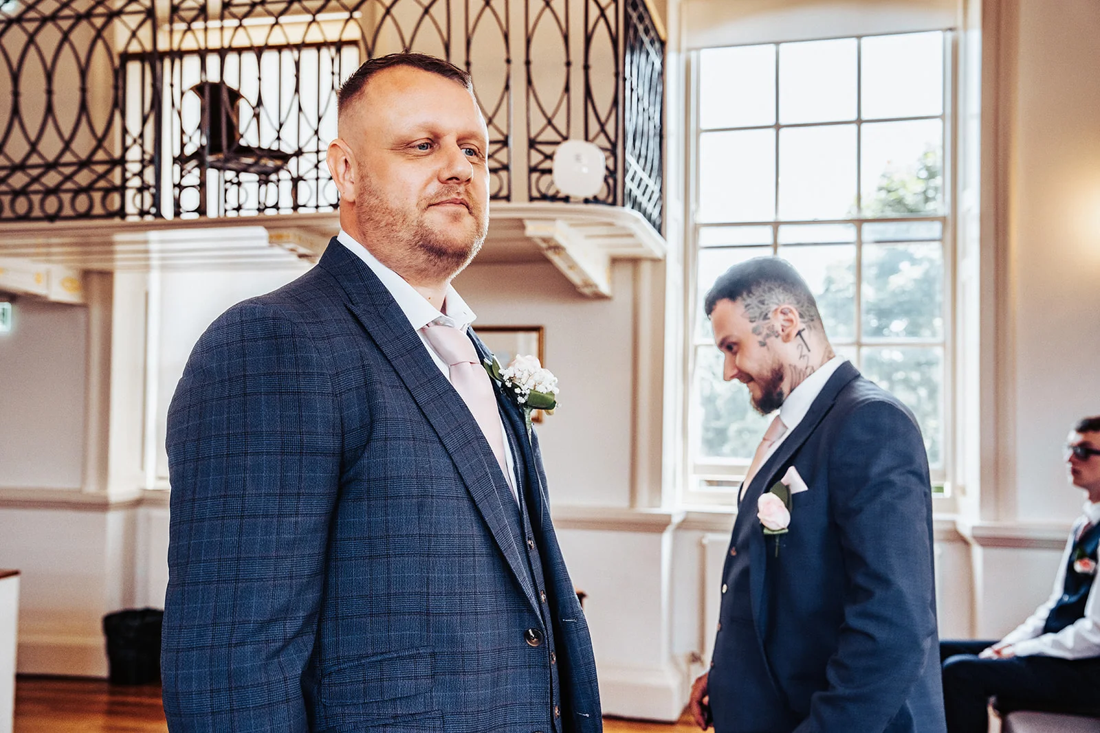 A tailored groom in a checked navy suit and pink tie waits at the altar inside a bright venue with large windows.