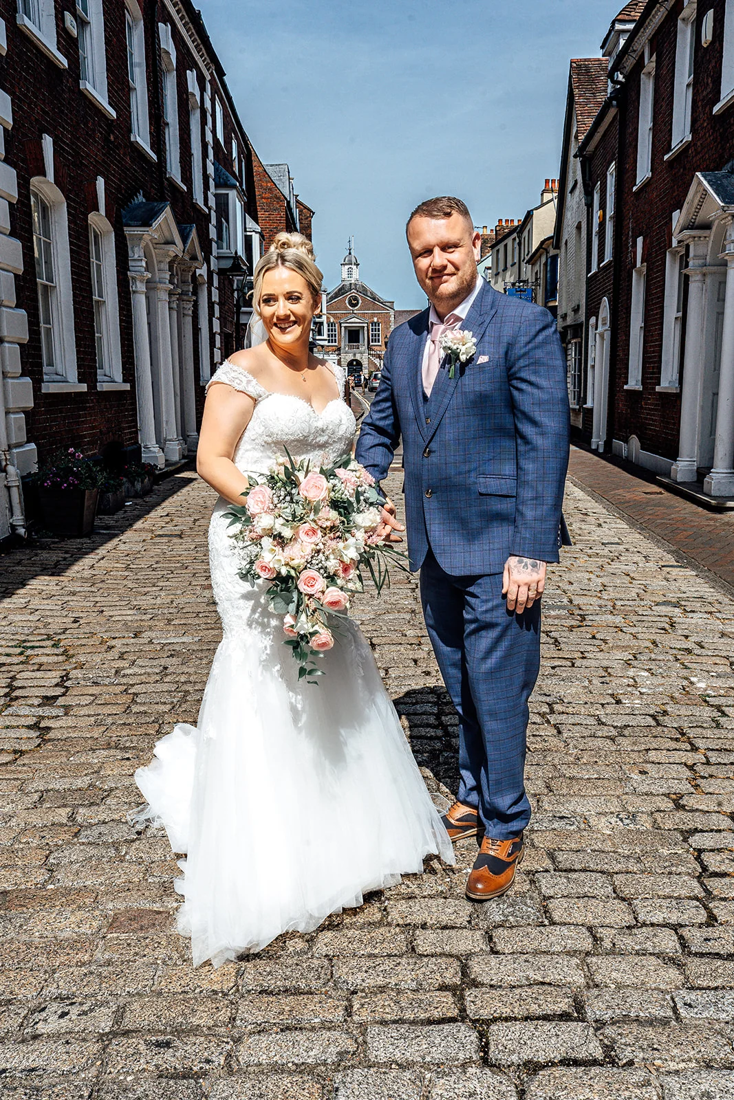 A bride in a lace fit-and-flare gown with a cascading floral bouquet stands arm-in-arm with a groom in a blue checked suit on a sunny cobbled street.