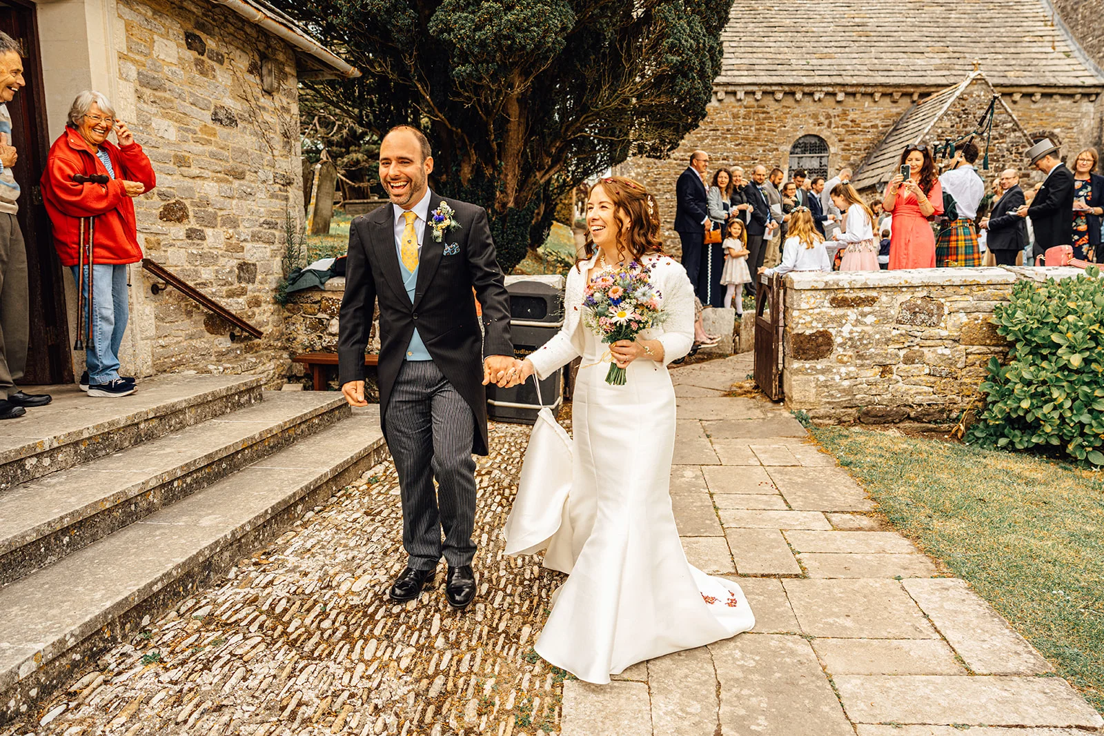 A joyous newlywed couple exit a historic stone church hand in hand, smiling broadly as wedding guests watch from the cobblestone pathway.