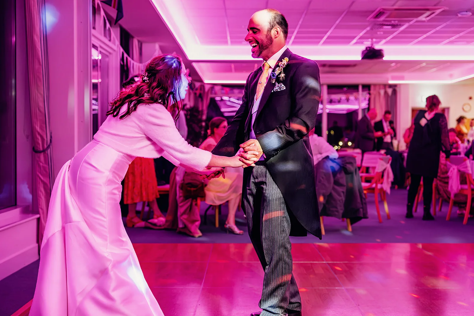A bride and groom share a joyful first dance together under vibrant pink lighting on a wooden dance floor at their evening reception.