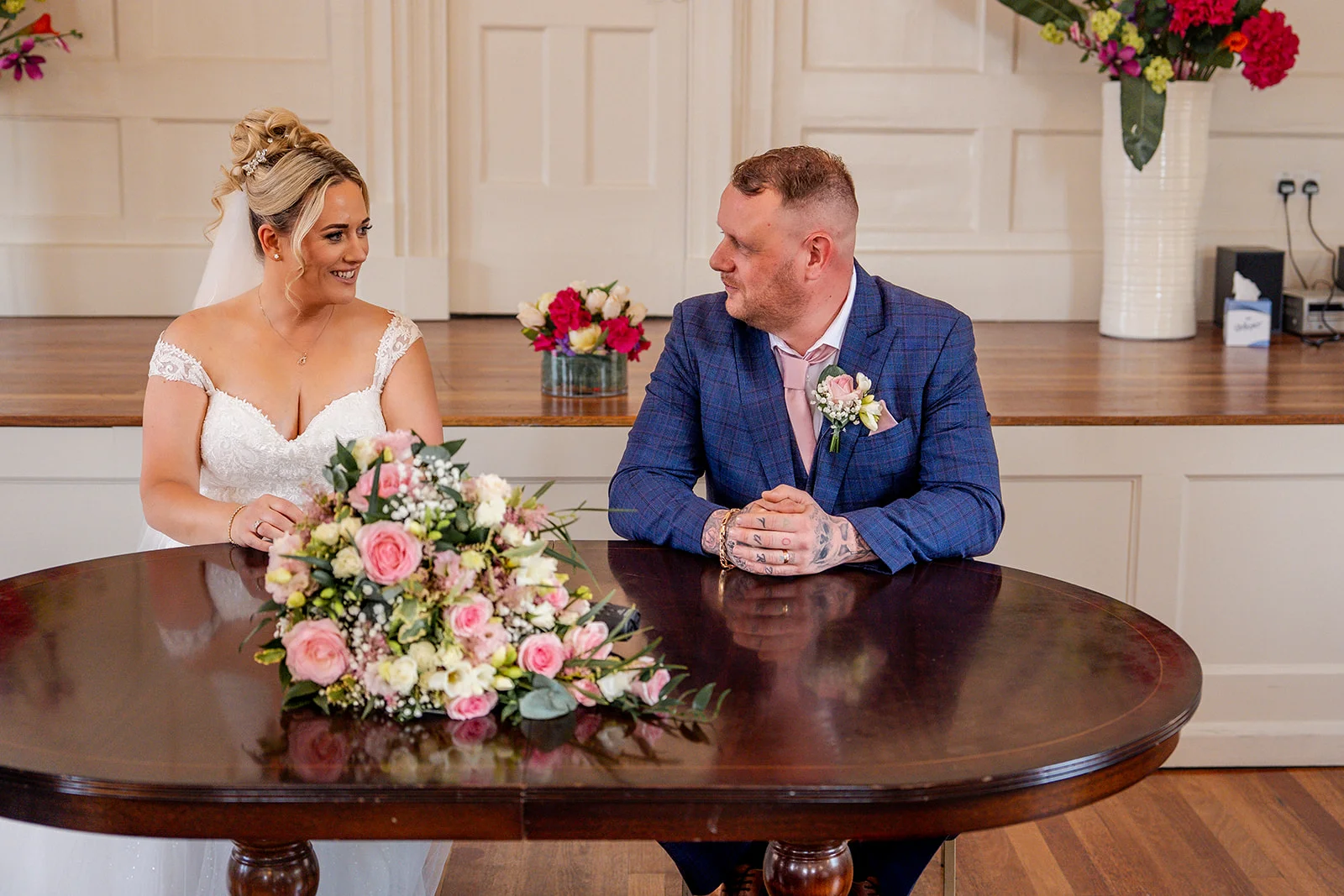 A bride and groom sit smiling at a dark wood table during their wedding ceremony, with a floral bouquet resting on the table in front of them.