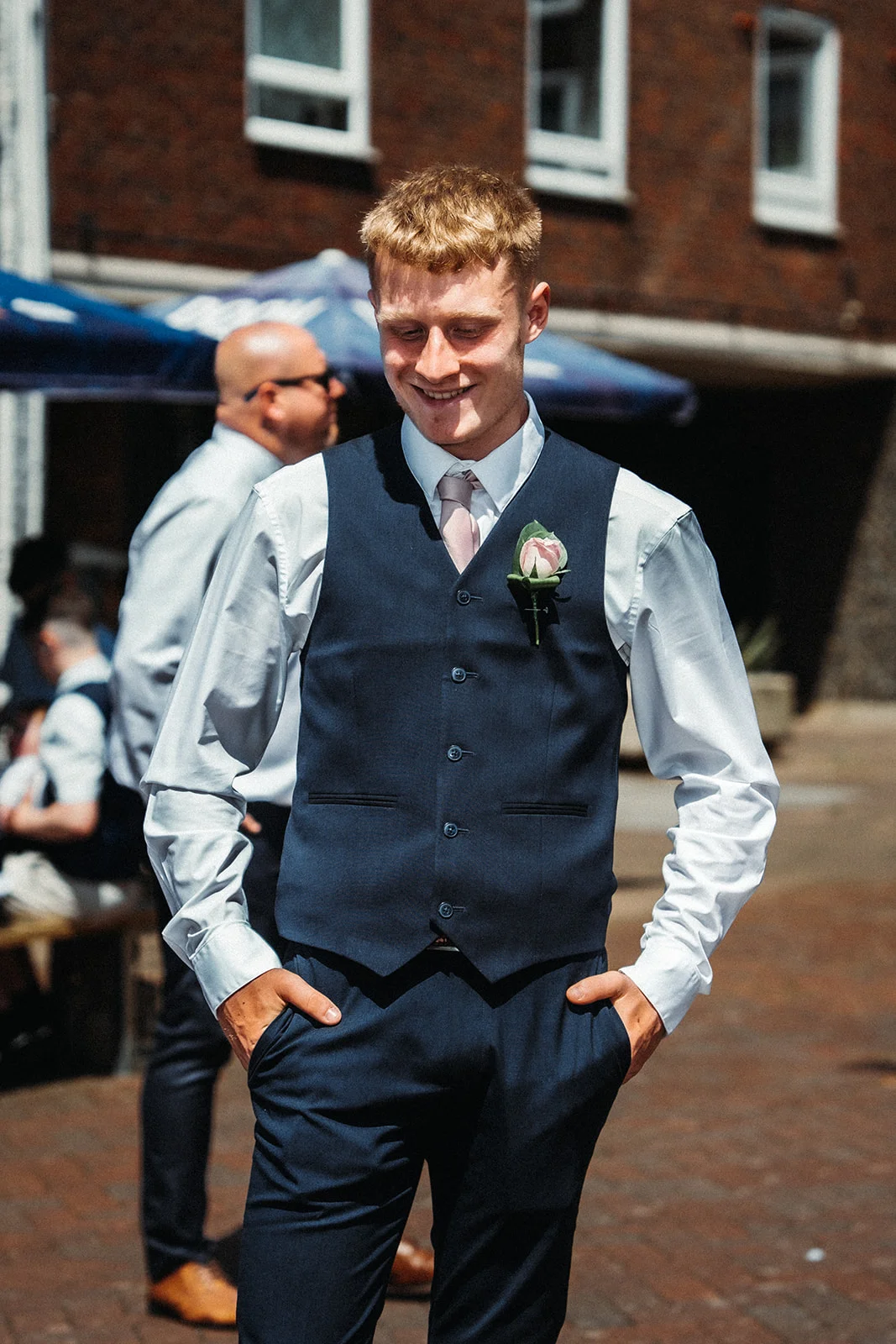 A smiling groom wearing a navy blue waistcoat and trousers with a pink rose buttonhole stands outdoors with his hands in his pockets.